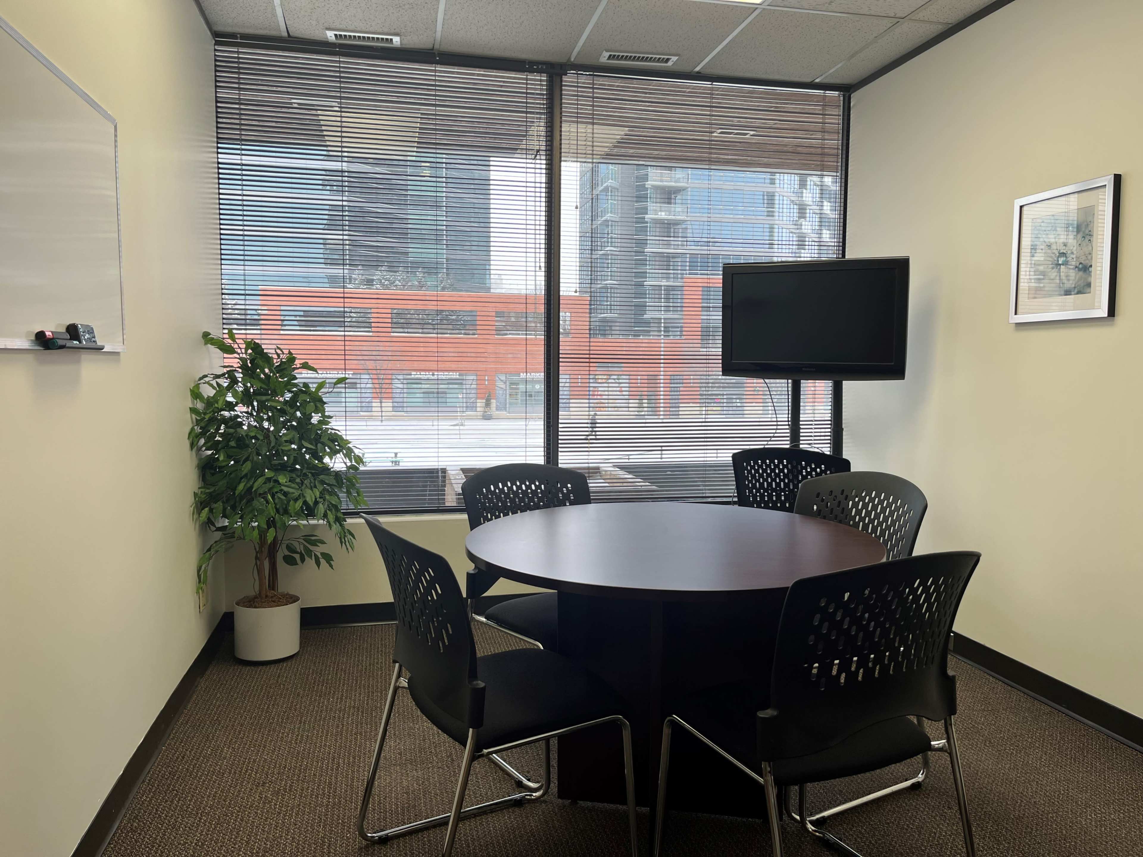 A small, modern conference room features a round table surrounded by four black chairs, with a television mounted on the wall and a large window overlooking a cityscape.