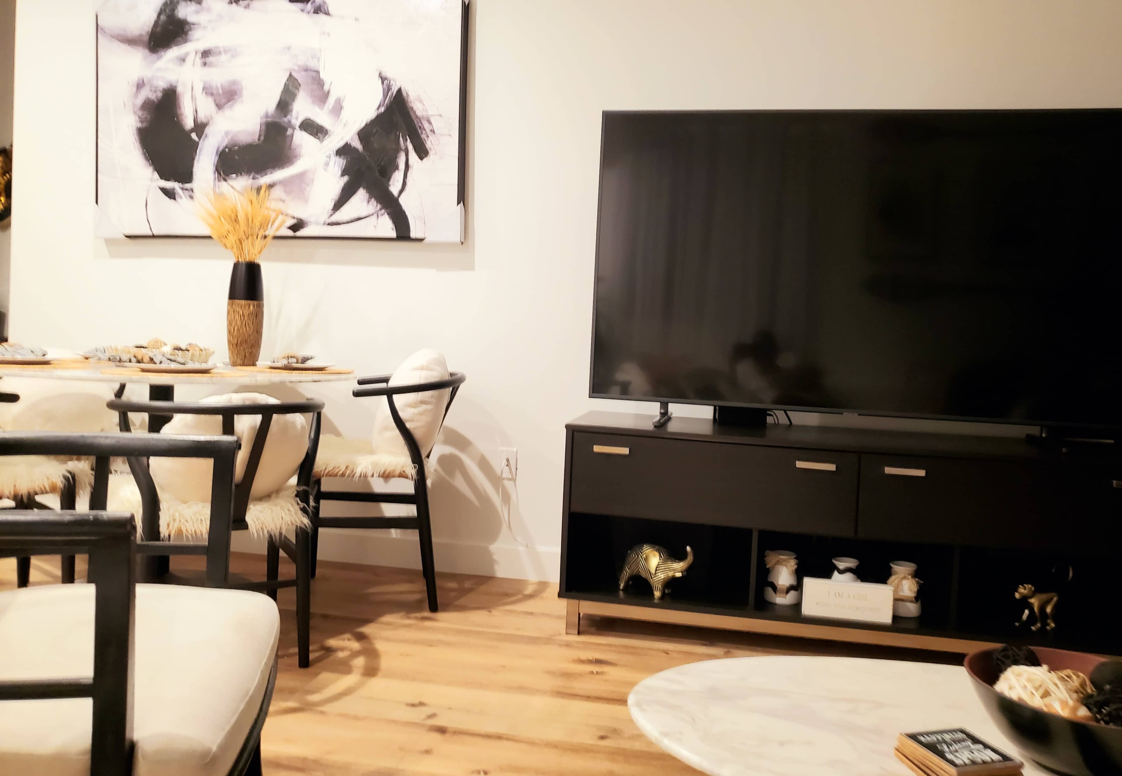 A modern living room features a television on a black media console, paired with a dining area that includes a table and chairs, along with abstract artwork on the wall.