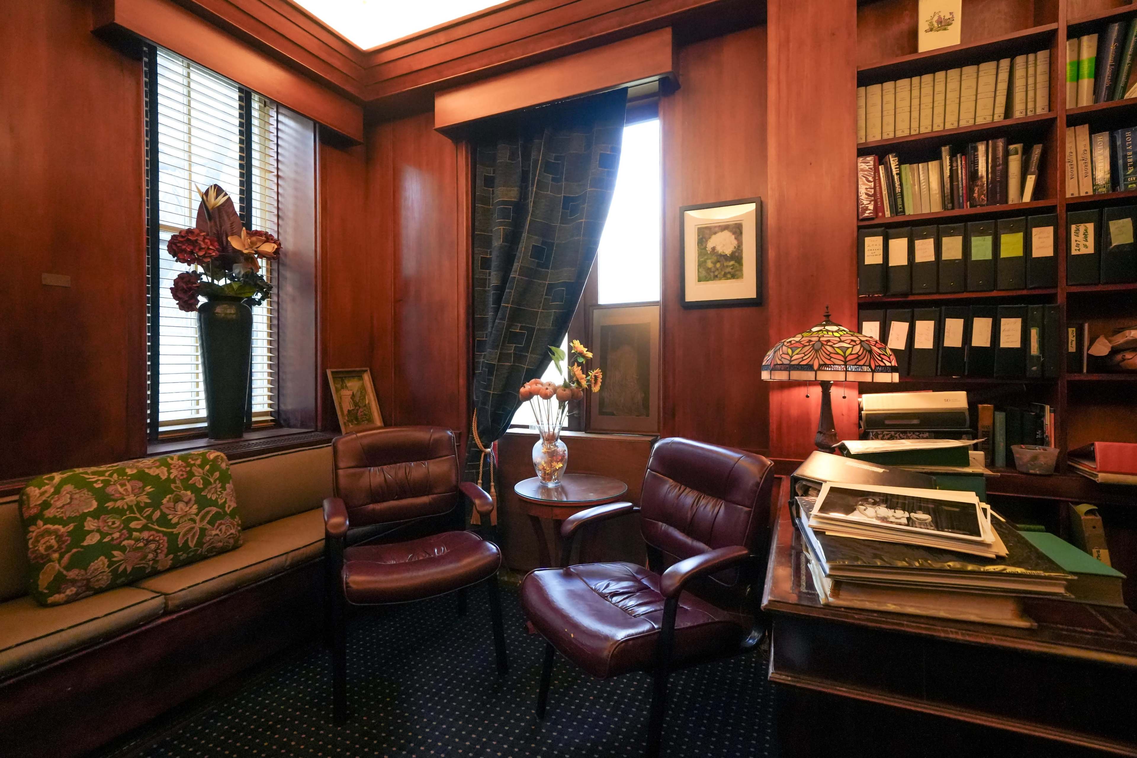 A cozy office space with two burgundy chairs, a wooden desk stacked with files, a stained glass lamp, and potted plants near a window.