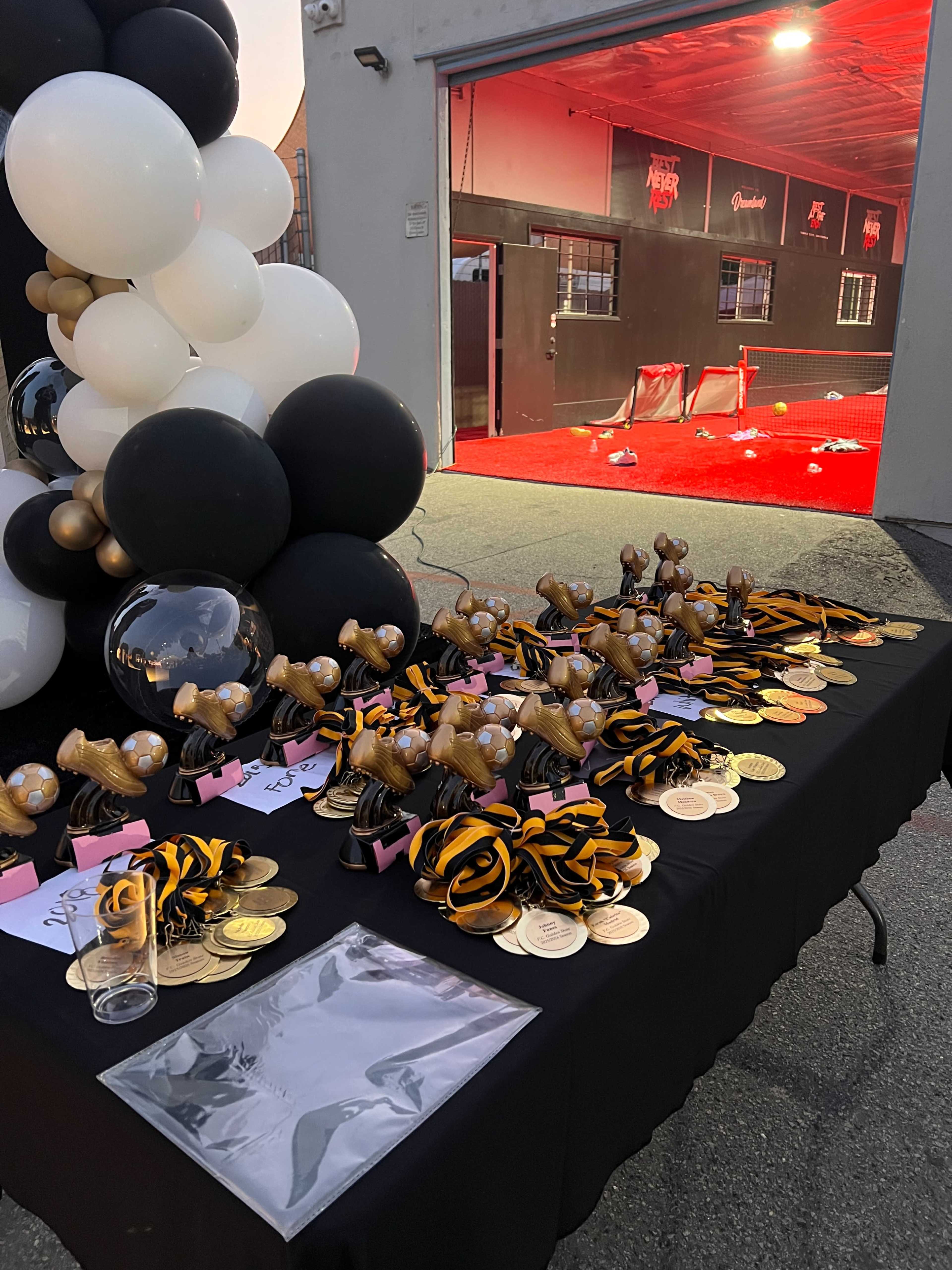 A table stacked with various trophies and medals, decorated with black and gold ribbons, is set up outside a building with red lights visible in the background.