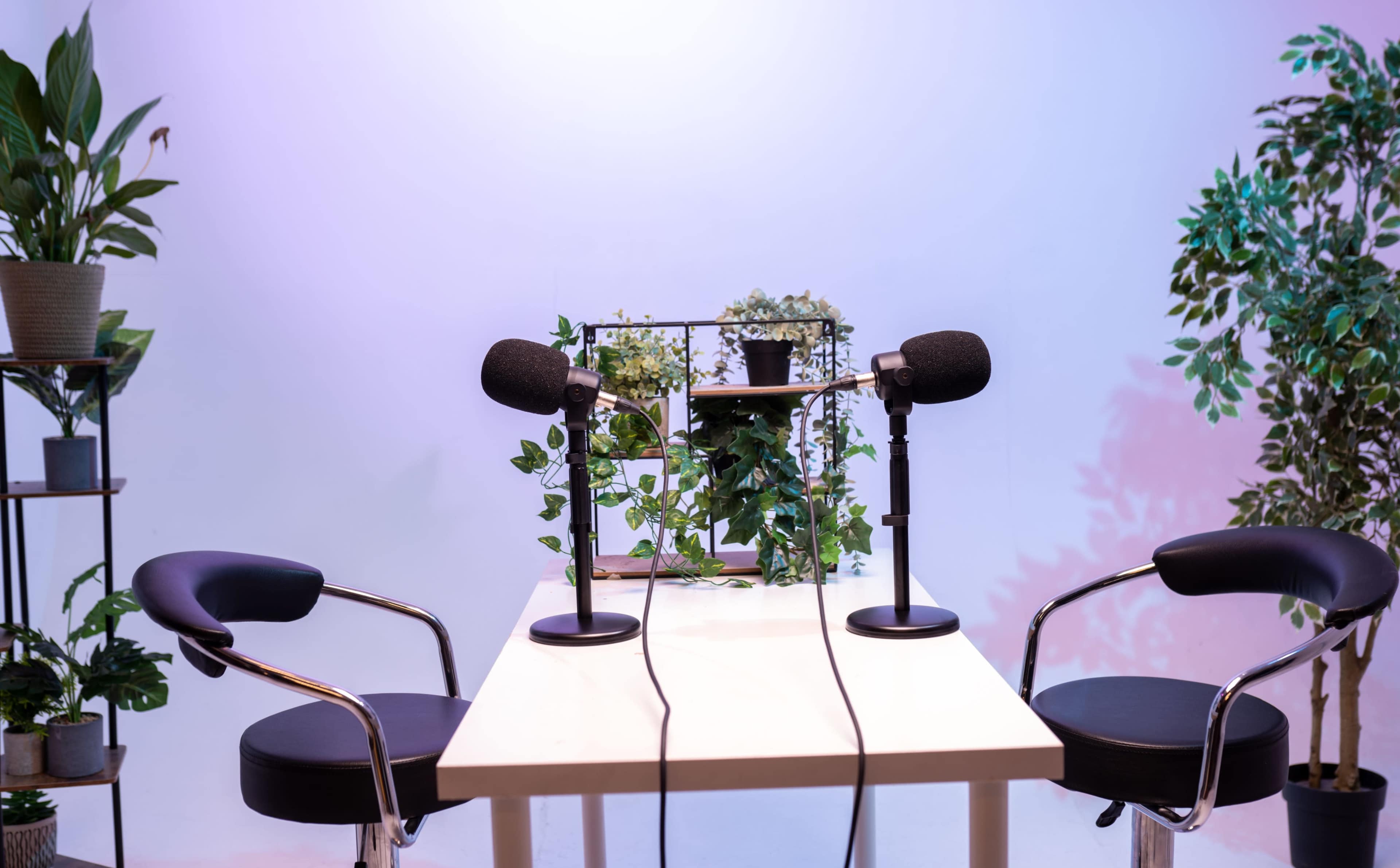 A small podcast studio features a table with two microphones and two chairs, surrounded by potted plants.