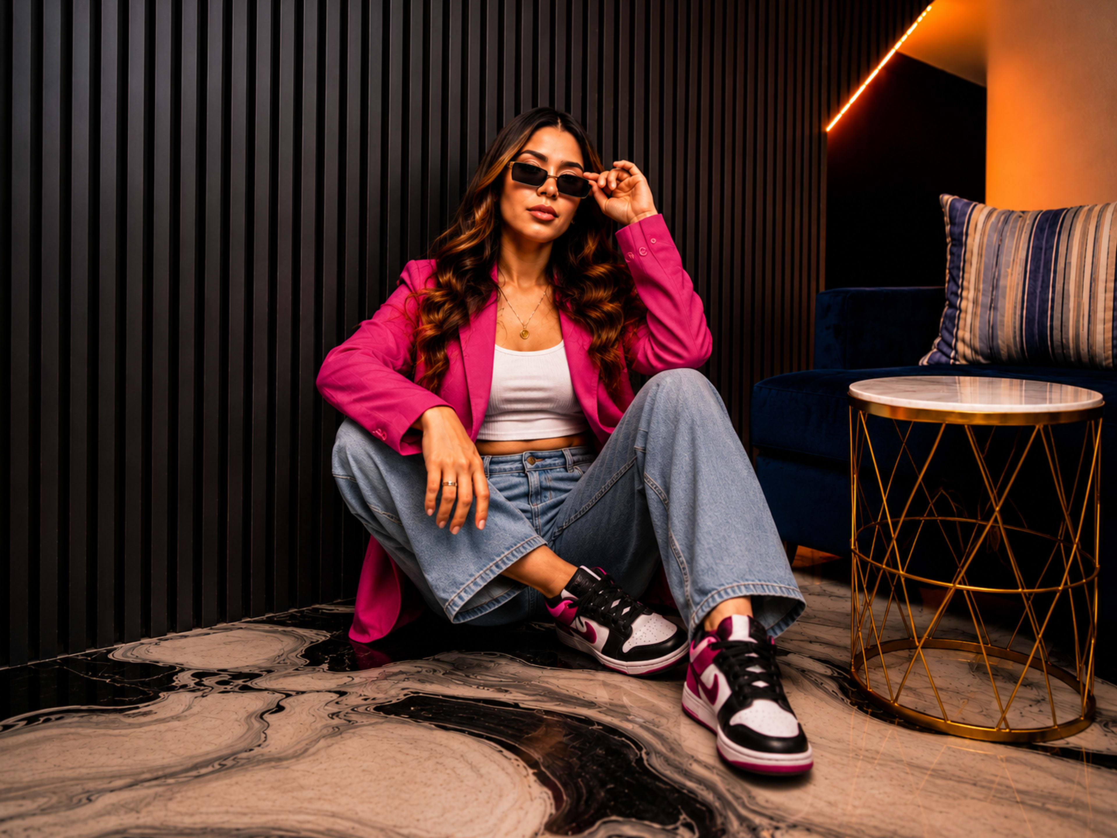 A woman in a pink blazer and sunglasses sits on the floor, wearing light blue jeans and sneakers, beside a gold side table in a modern interior.