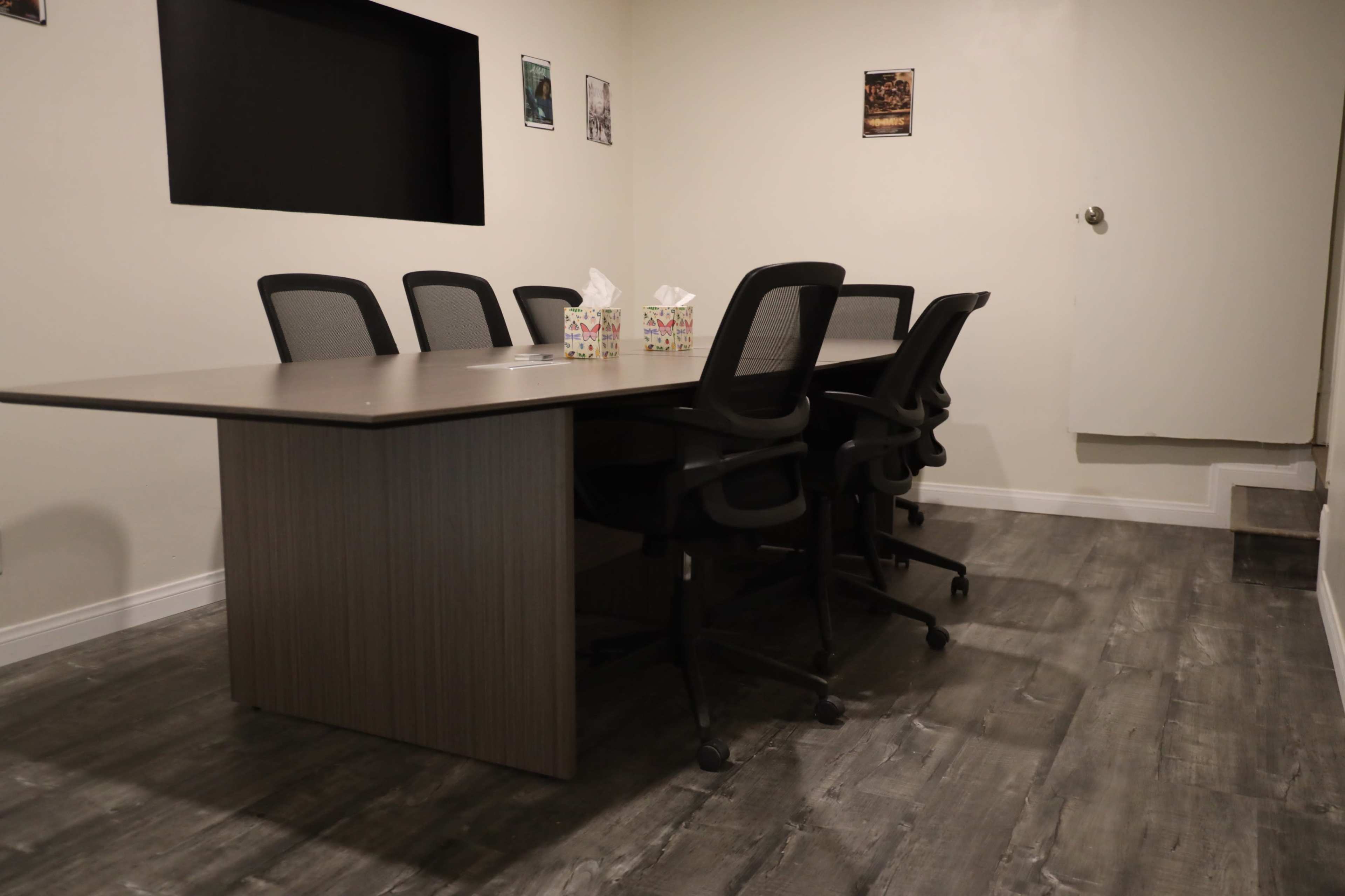 The image shows a conference room with a large table surrounded by several ergonomic chairs and a wall featuring a black screen and framed pictures.