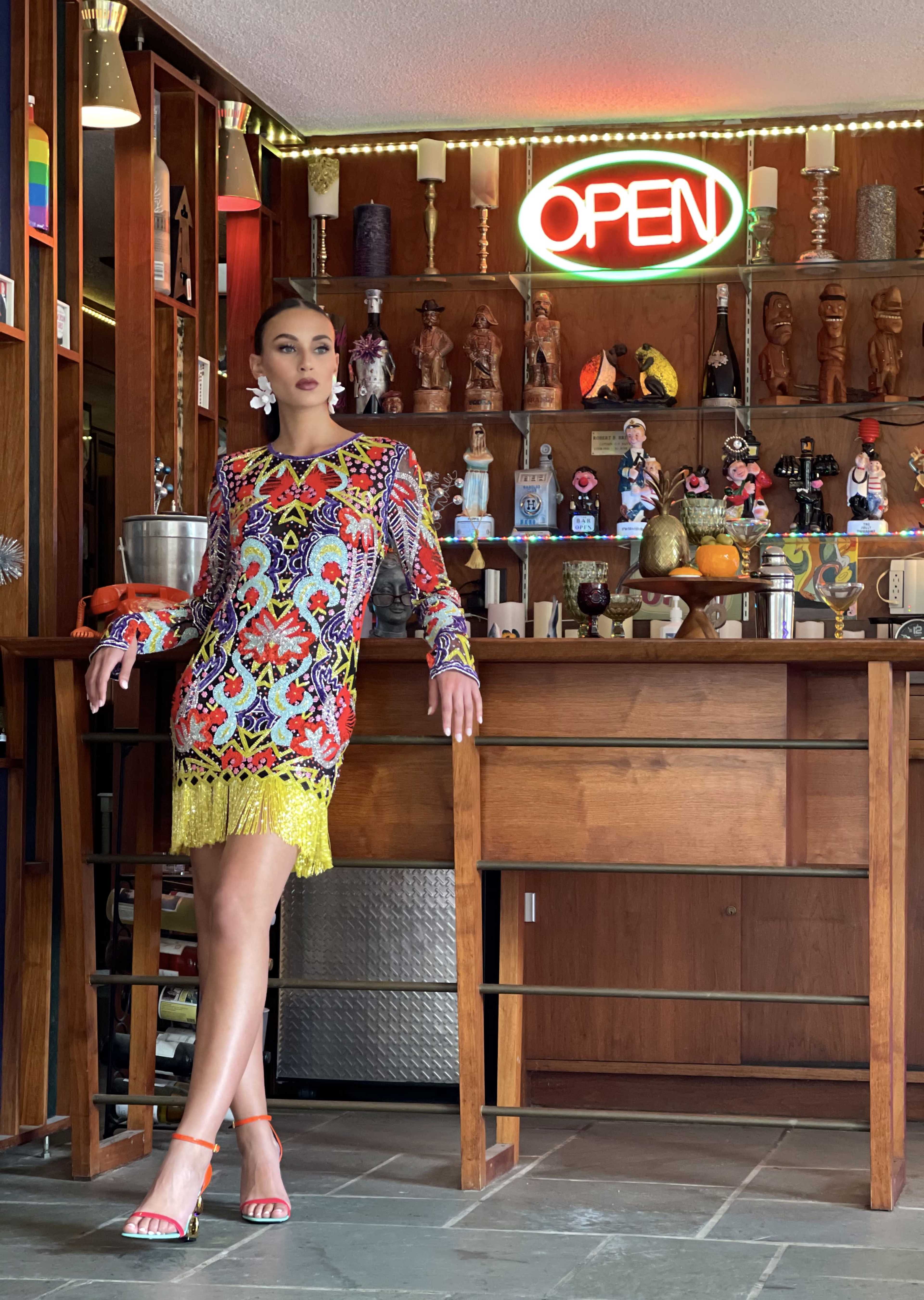 A woman poses in a colorful patterned dress with fringe against a backdrop of a well-decorated bar featuring various collectibles and an illuminated "OPEN" sign.