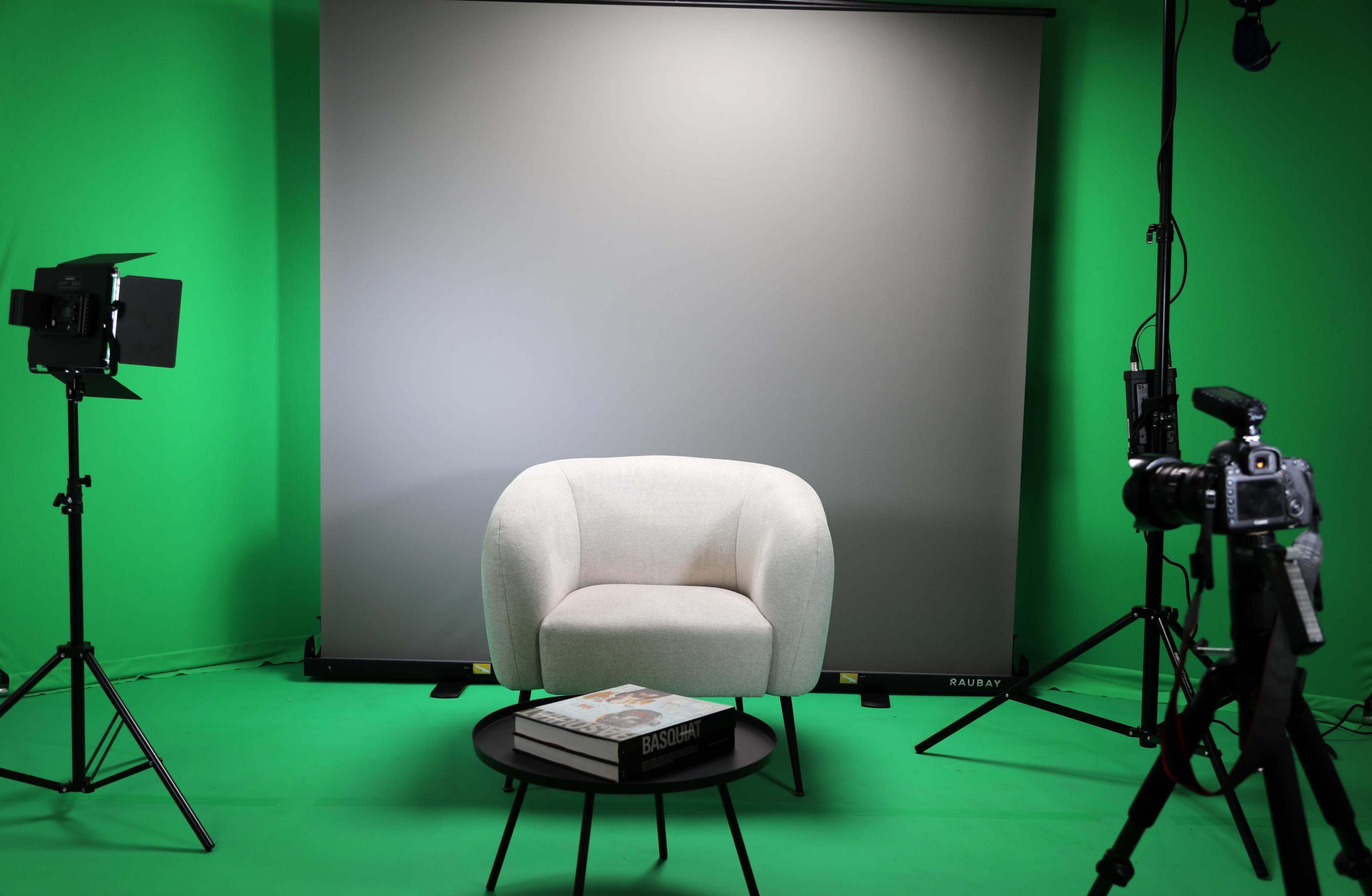 A circular white armchair is positioned in front of a gray backdrop, with a small black coffee table holding a book and cameras set up on tripods nearby.