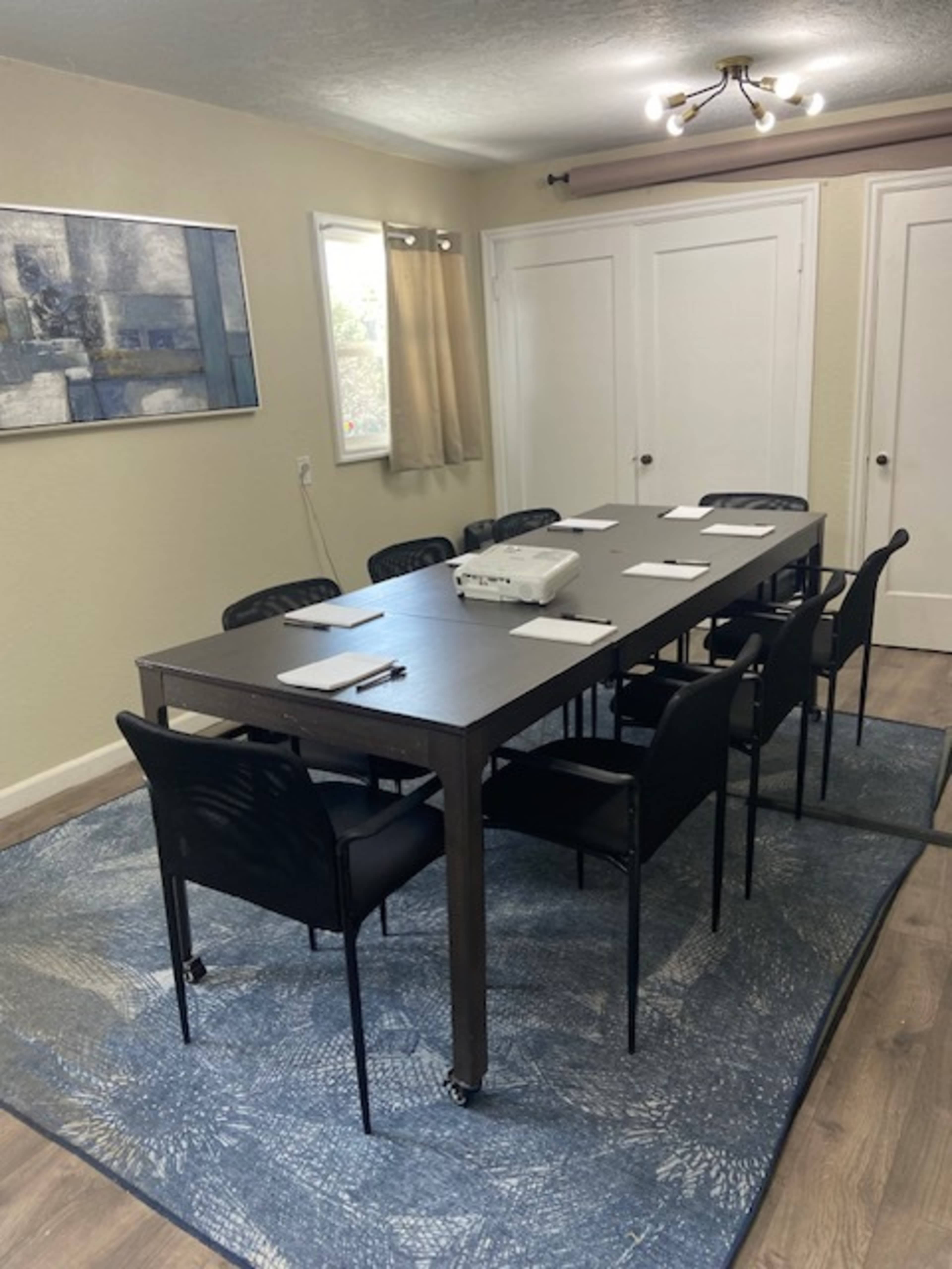 A rectangular conference table with eight black chairs is arranged on a blue rug in a well-lit room with two doors and a wall-mounted projector.