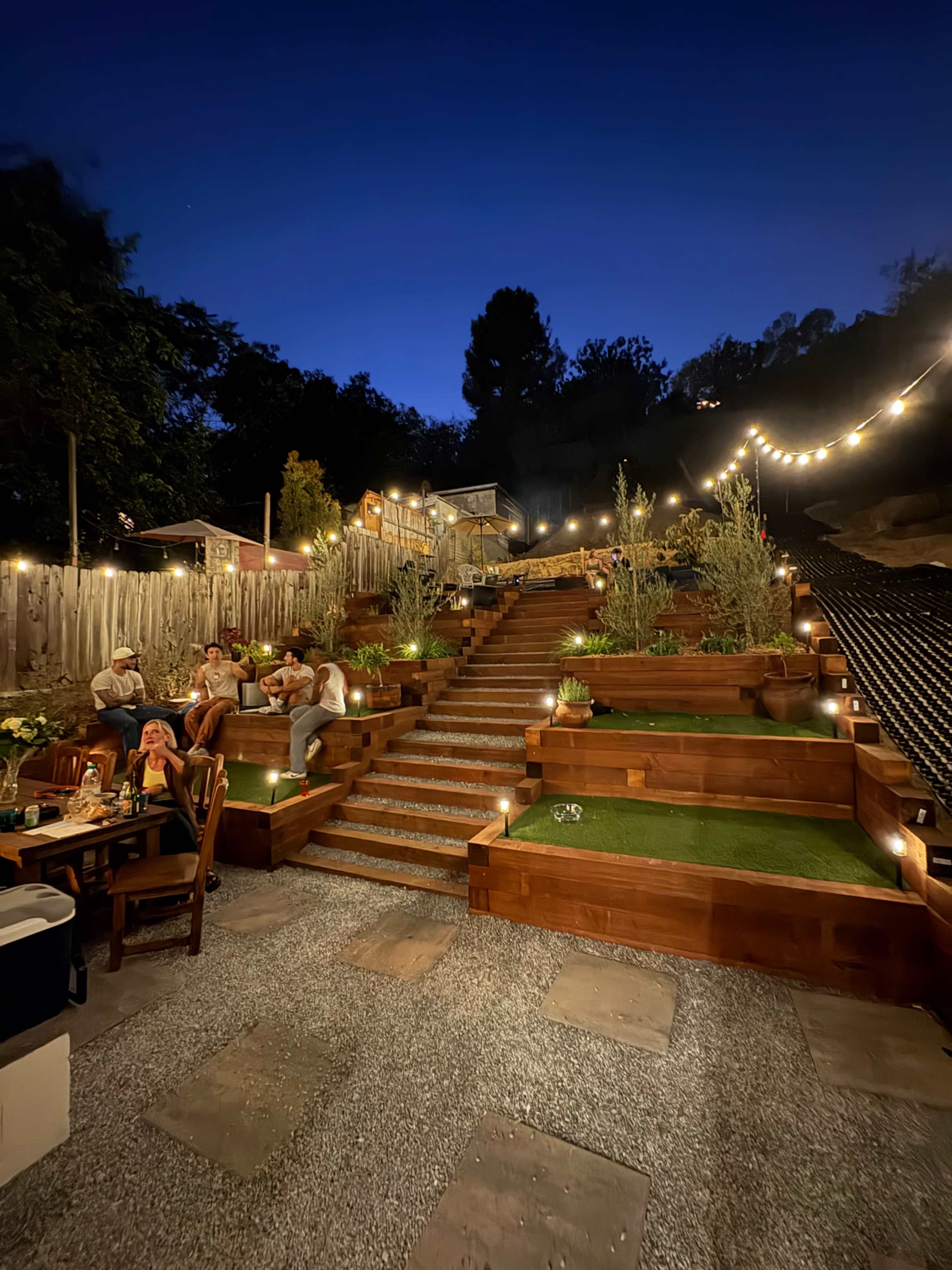A multi-level wooden terrace garden is illuminated with soft lights, featuring guests gathered on the steps and at a dining table in the foreground.