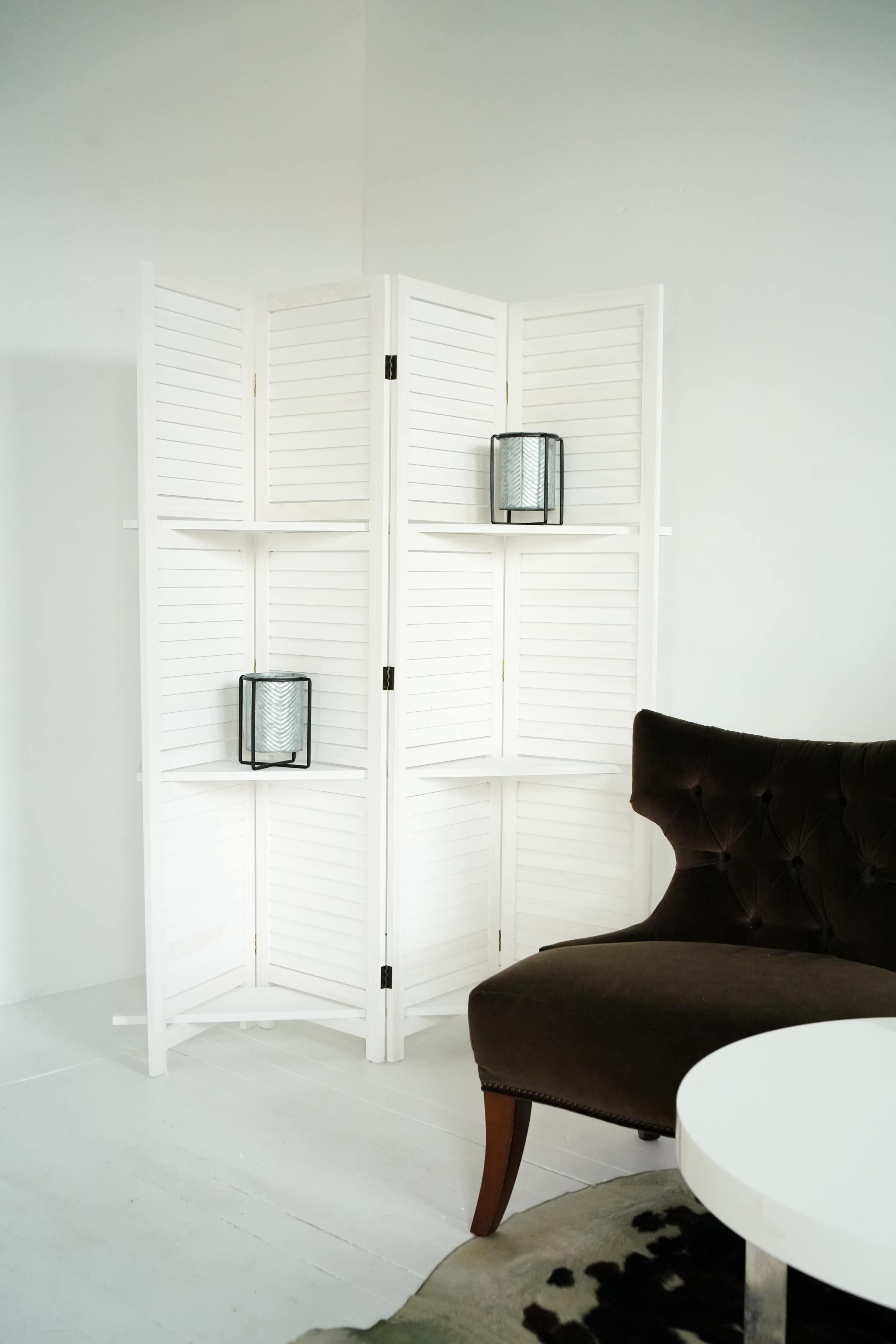 A white folding screen with shelves and two lanterns is positioned next to a dark upholstered chair and a round table in a minimalistic room.