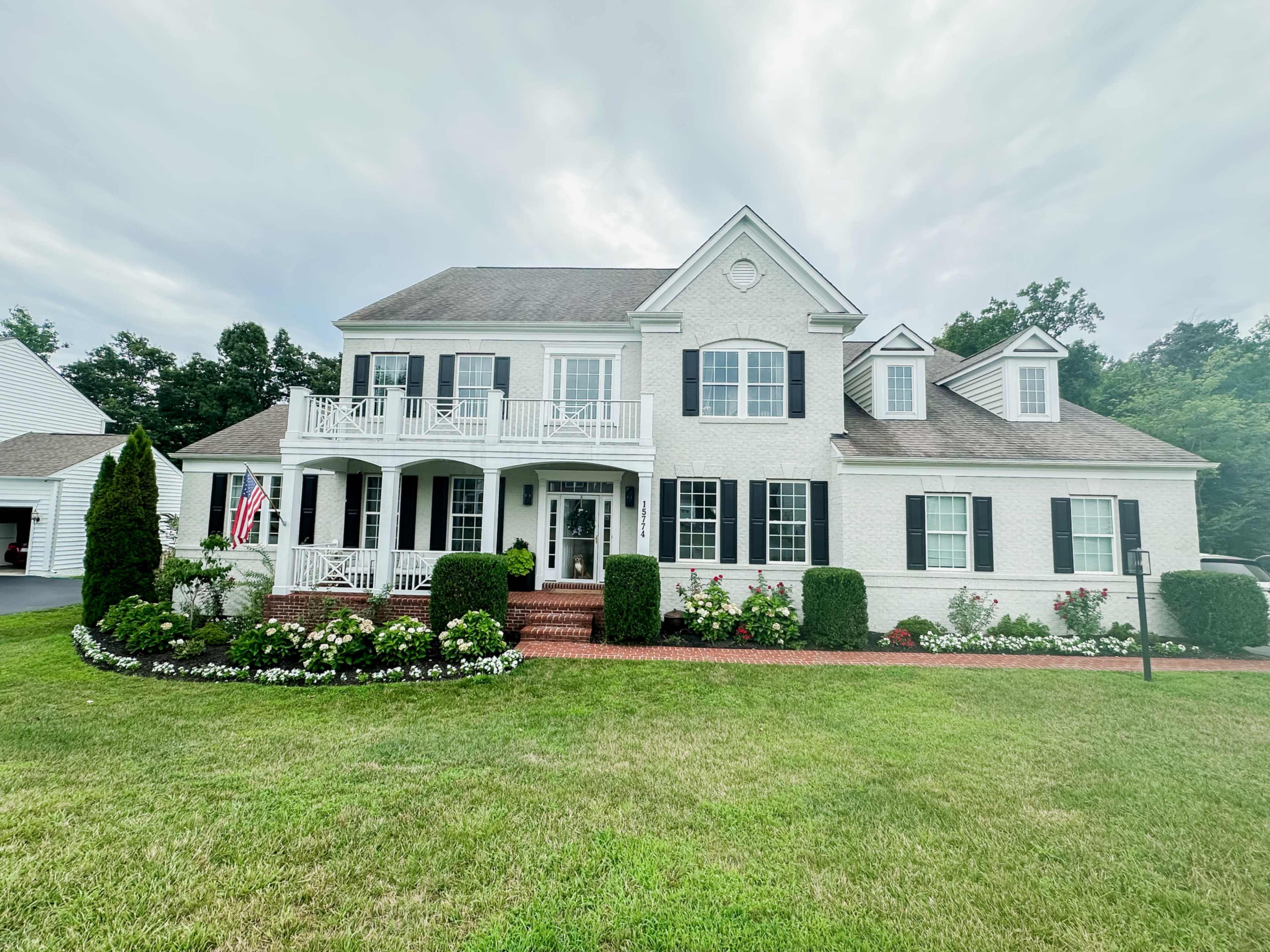 A two-story white house with black shutters, a brick walkway, and a landscaped front yard featuring flowers and a flagpole.