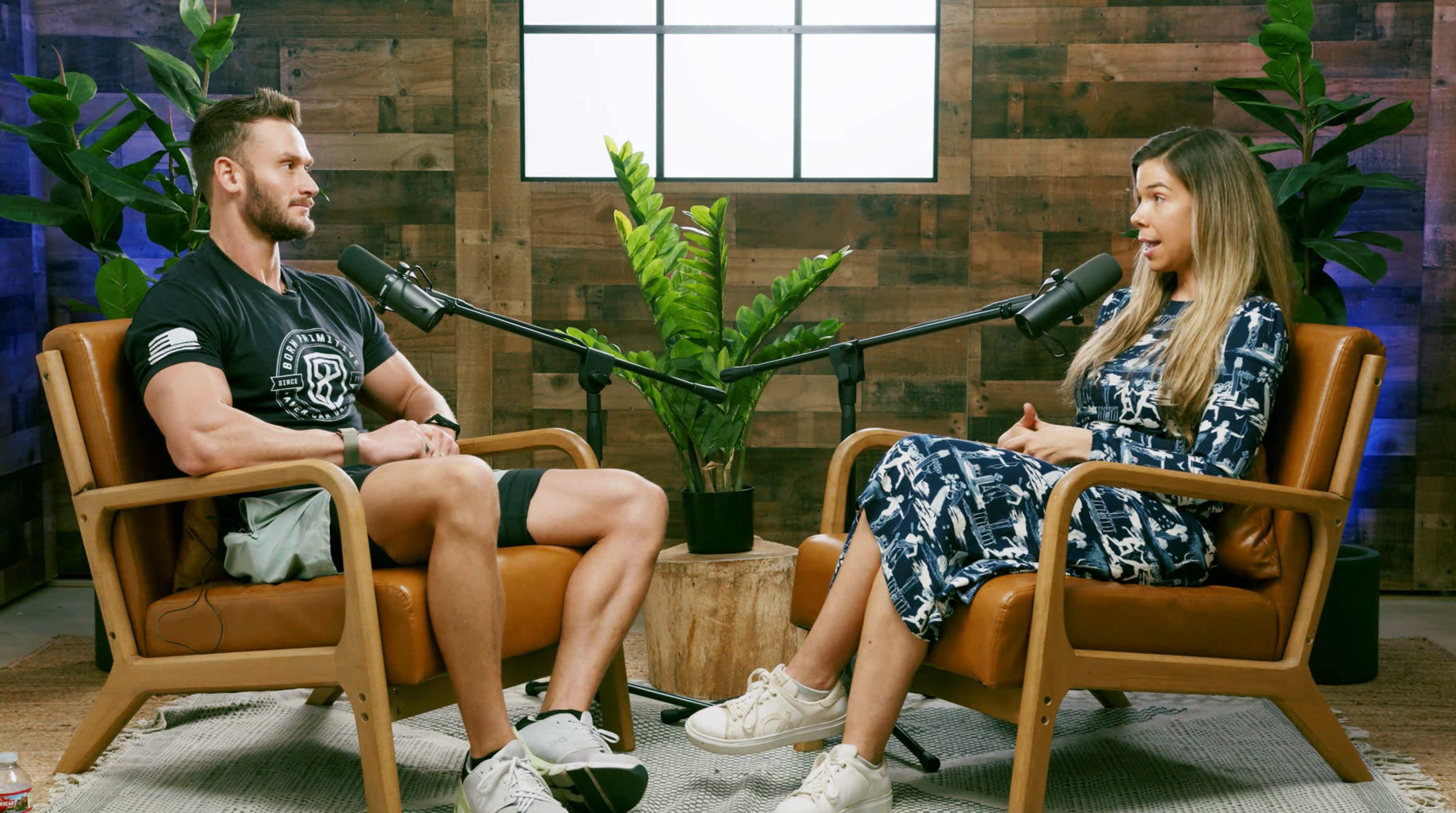A man and a woman sit in modern chairs facing each other in a room with wooden walls and a large window, both speaking into microphones.