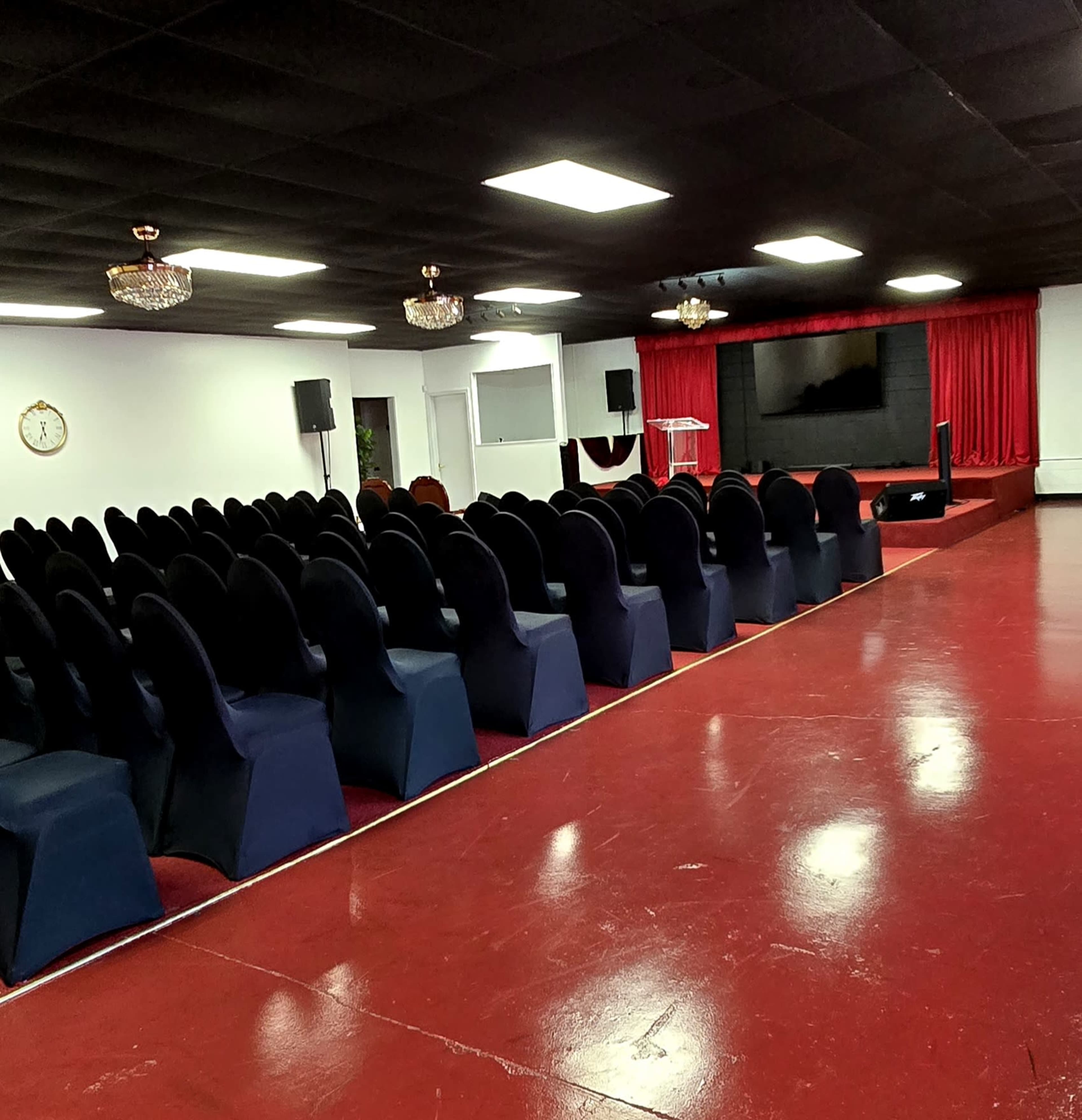 The image shows a spacious indoor auditorium with rows of black chairs facing a stage, which is illuminated by overhead lights and features red curtains and a large screen.