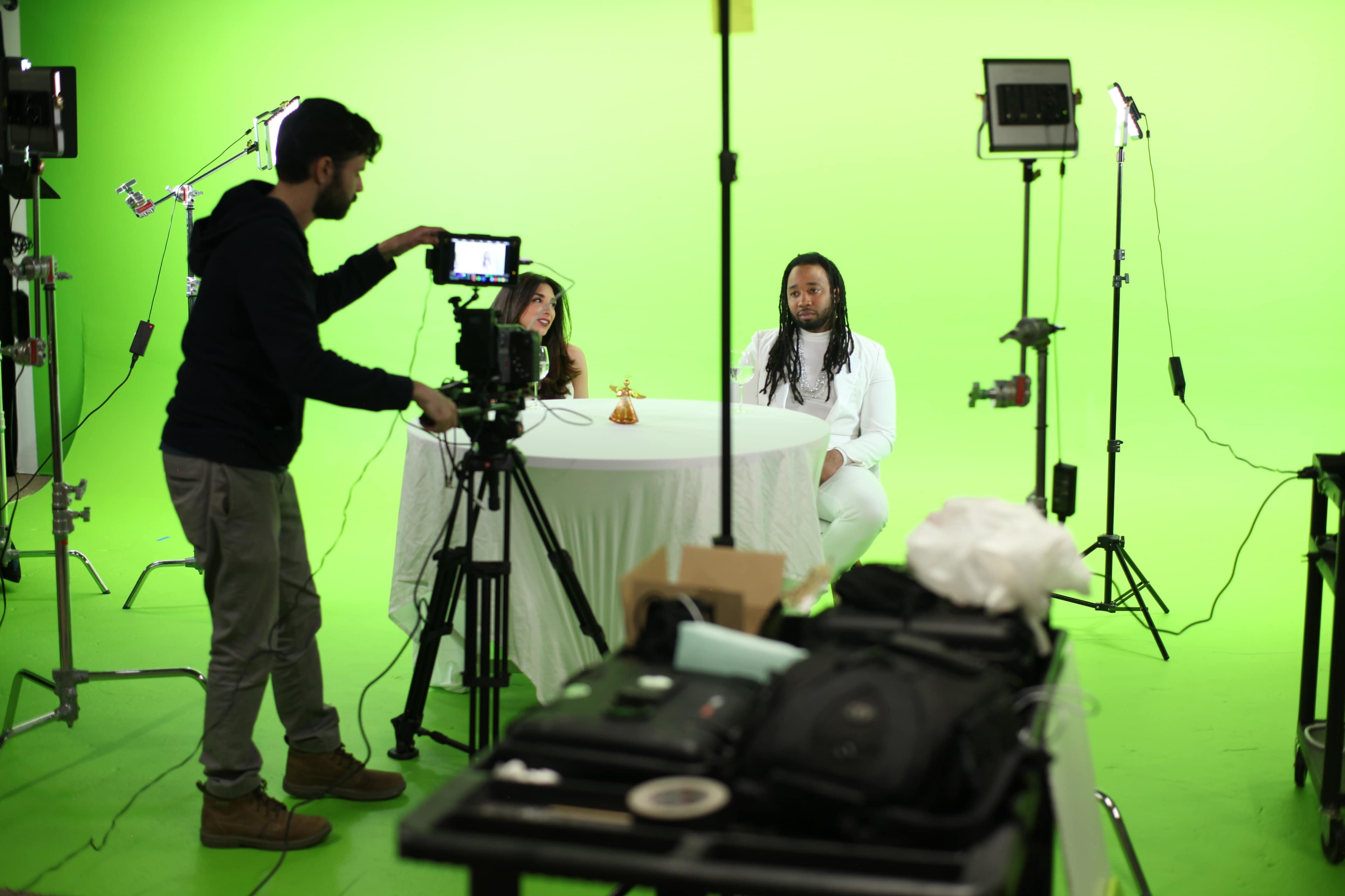 A man operates a camera while a woman and a man sit at a white table against a green screen backdrop.