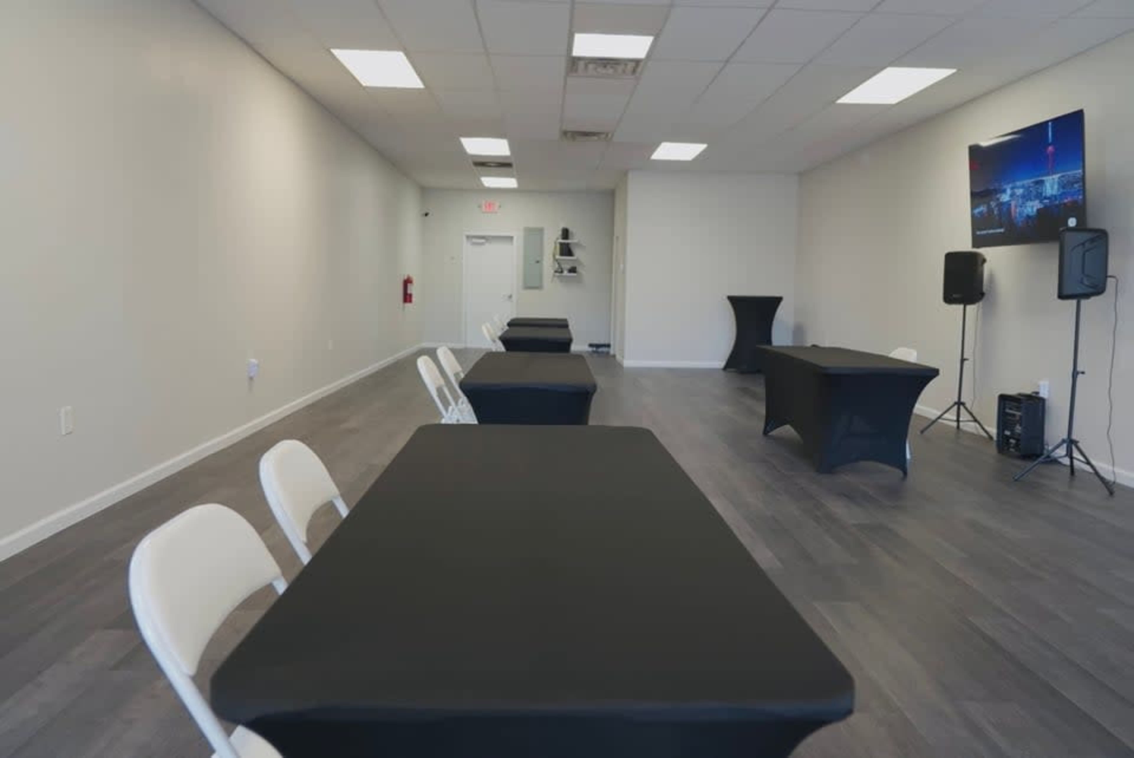 The image shows a spacious, empty room with several black tables set up, white chairs arranged neatly, and a television mounted on the wall.