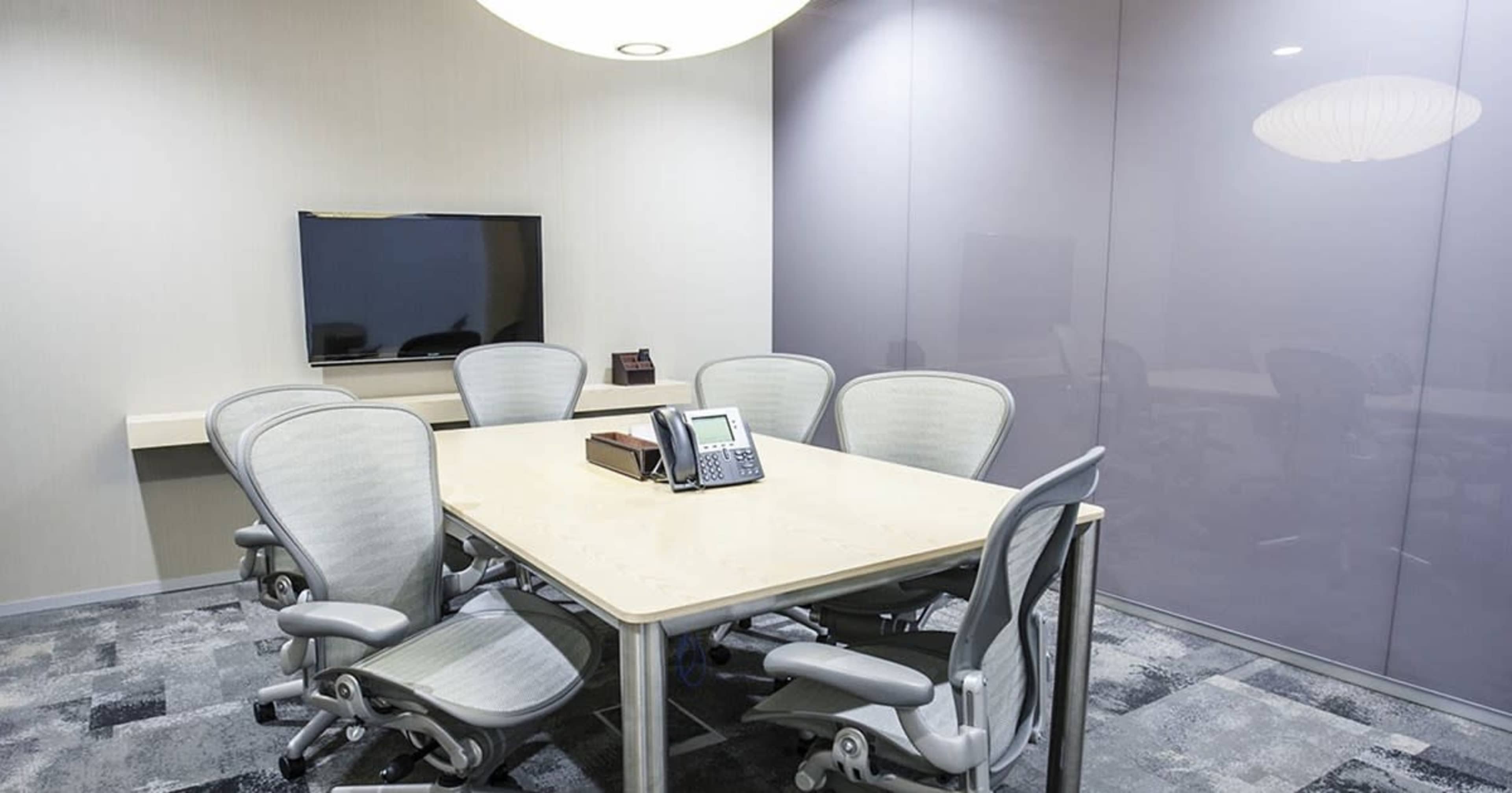 A conference room features a large wooden table surrounded by several ergonomic chairs, with a television mounted on the wall.