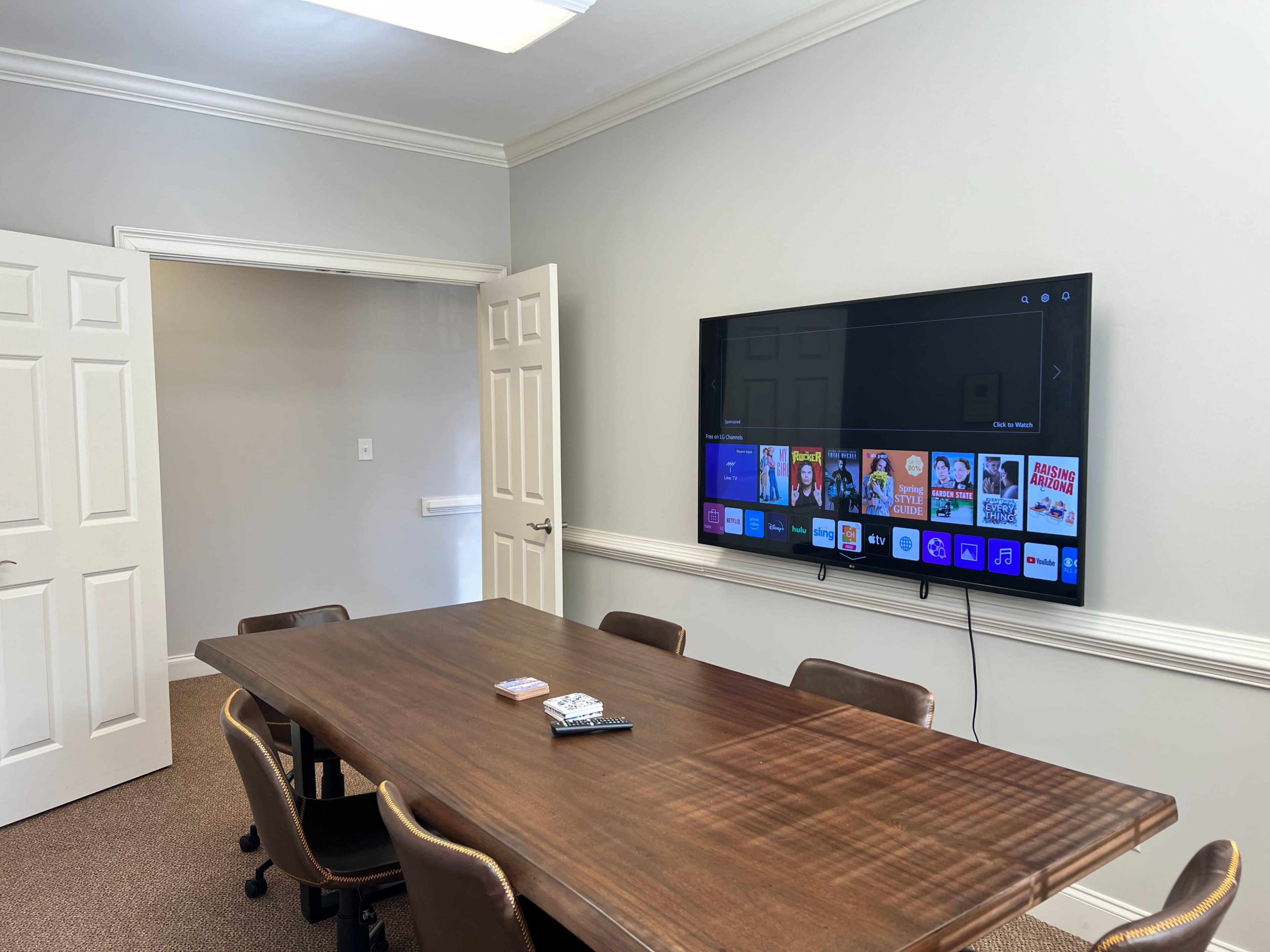 A conference room features a large wooden table with chairs and a wall-mounted television displaying a menu interface.