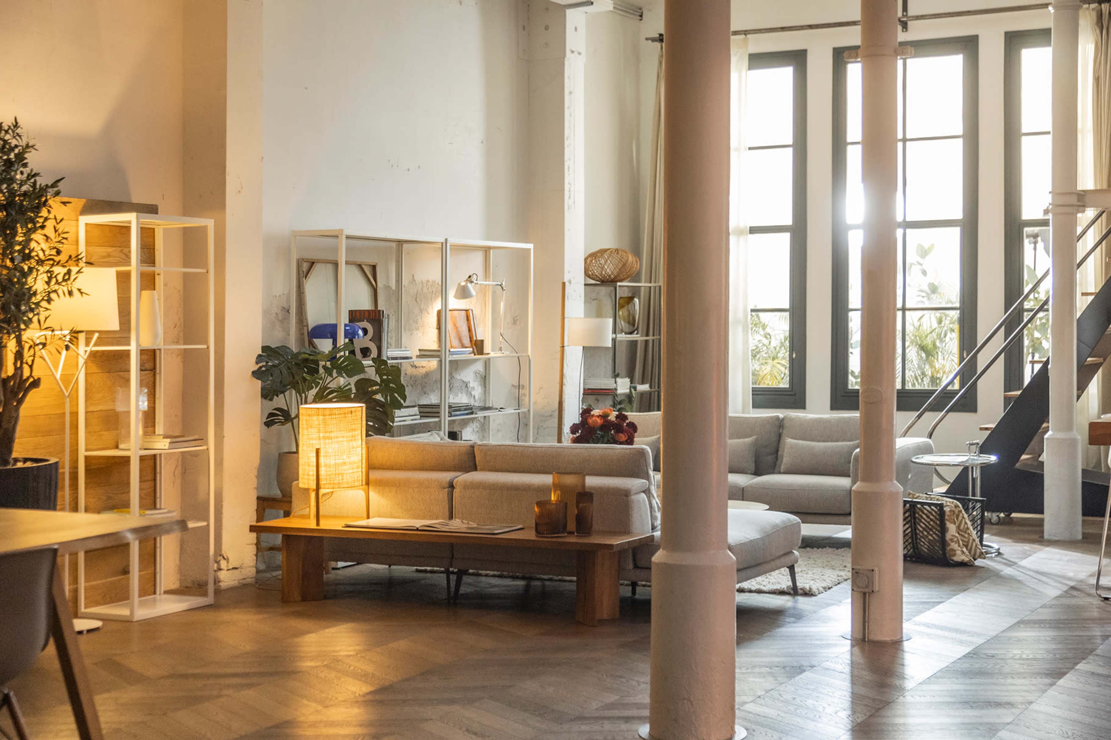 The image shows a spacious, well-lit living room with modern furniture, including a gray sectional sofa and wooden shelves.