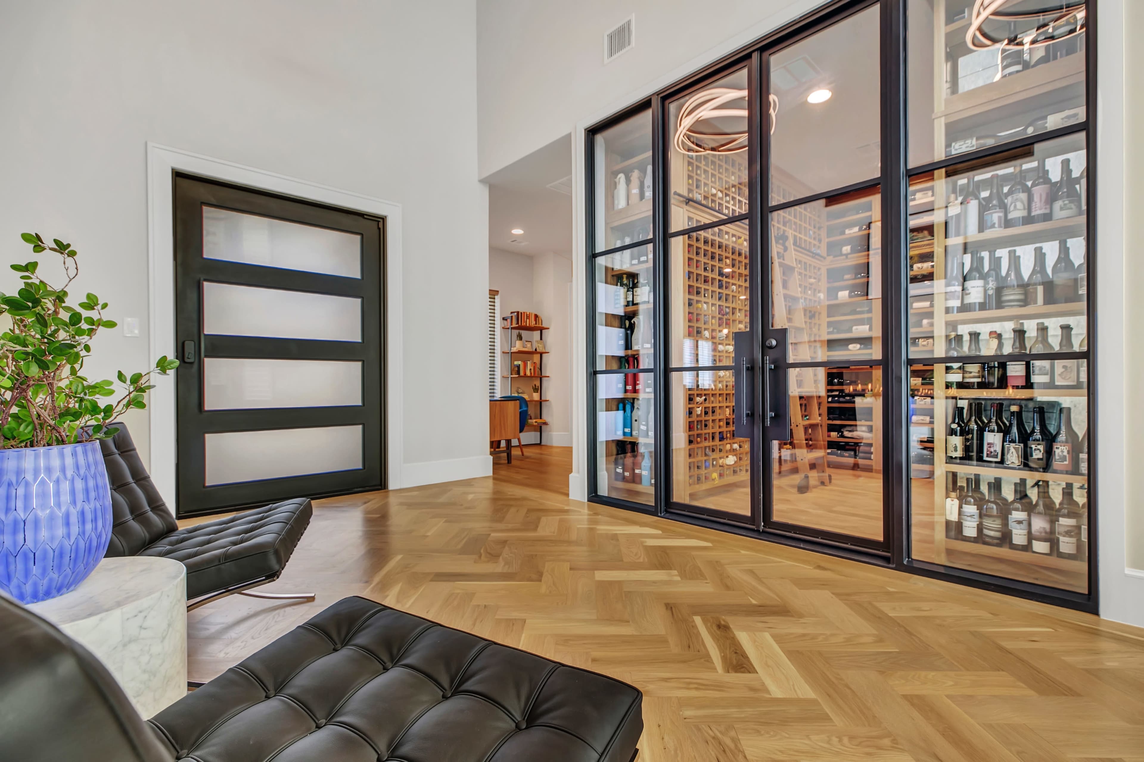 A modern interior with a glass-walled wine cellar, sleek furniture, and a large door, all set against a light hardwood floor.