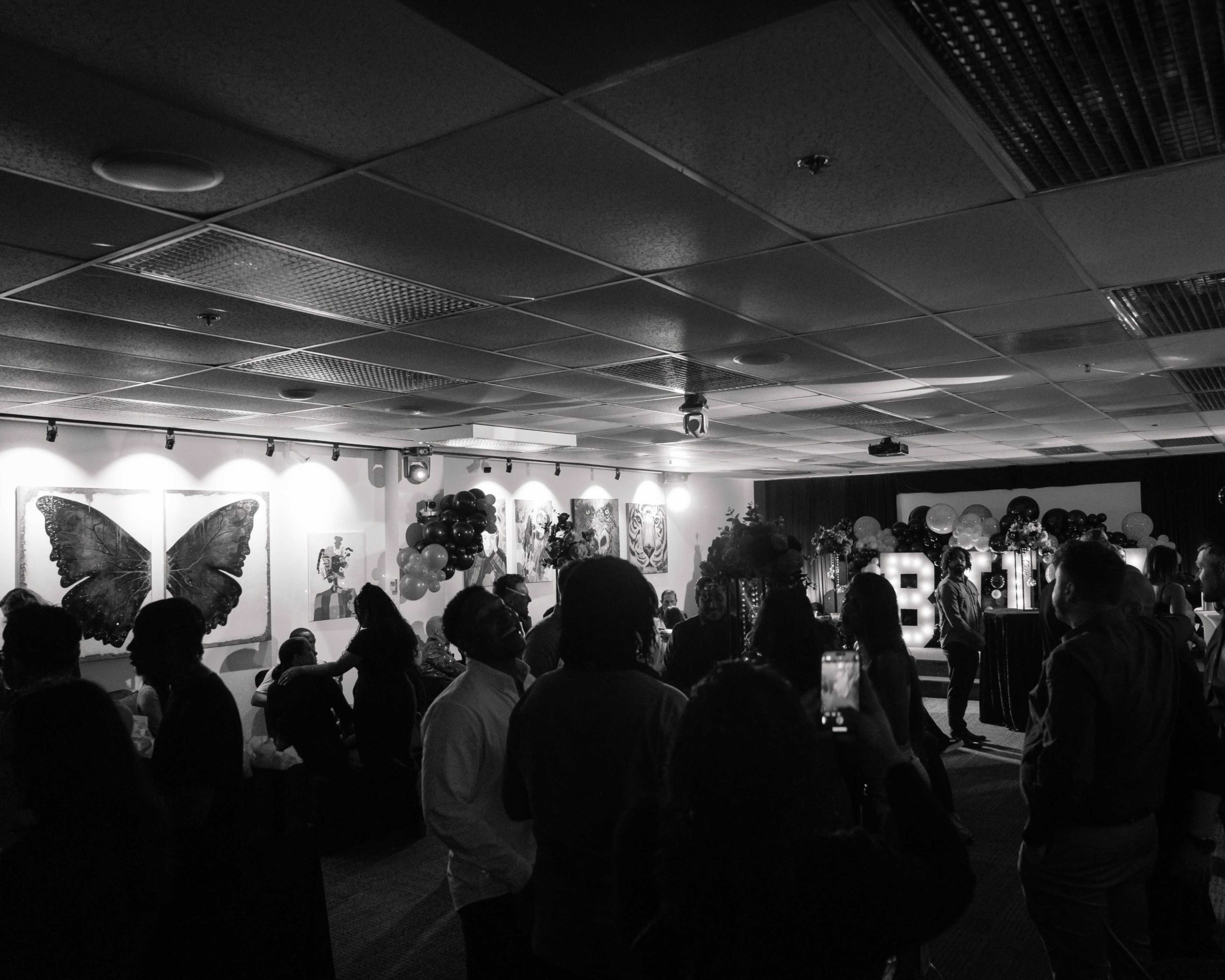 A crowd gathers in a dimly lit room adorned with artwork and balloons, with a large illuminated display spelling "BIRTHDAY" in the background.