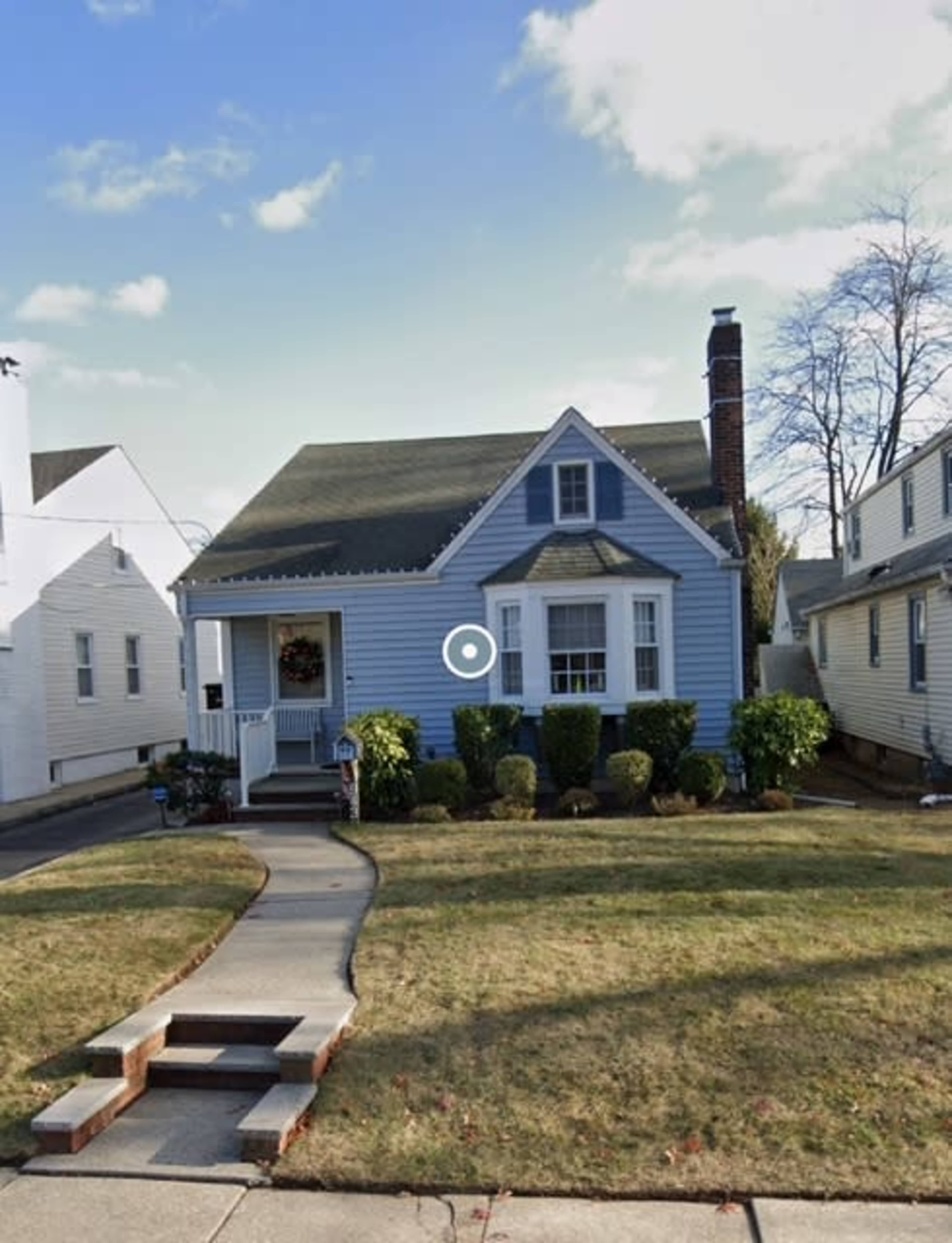 A small blue house with a peaked roof and a front walkway bordered by bushes is set on a suburban street.