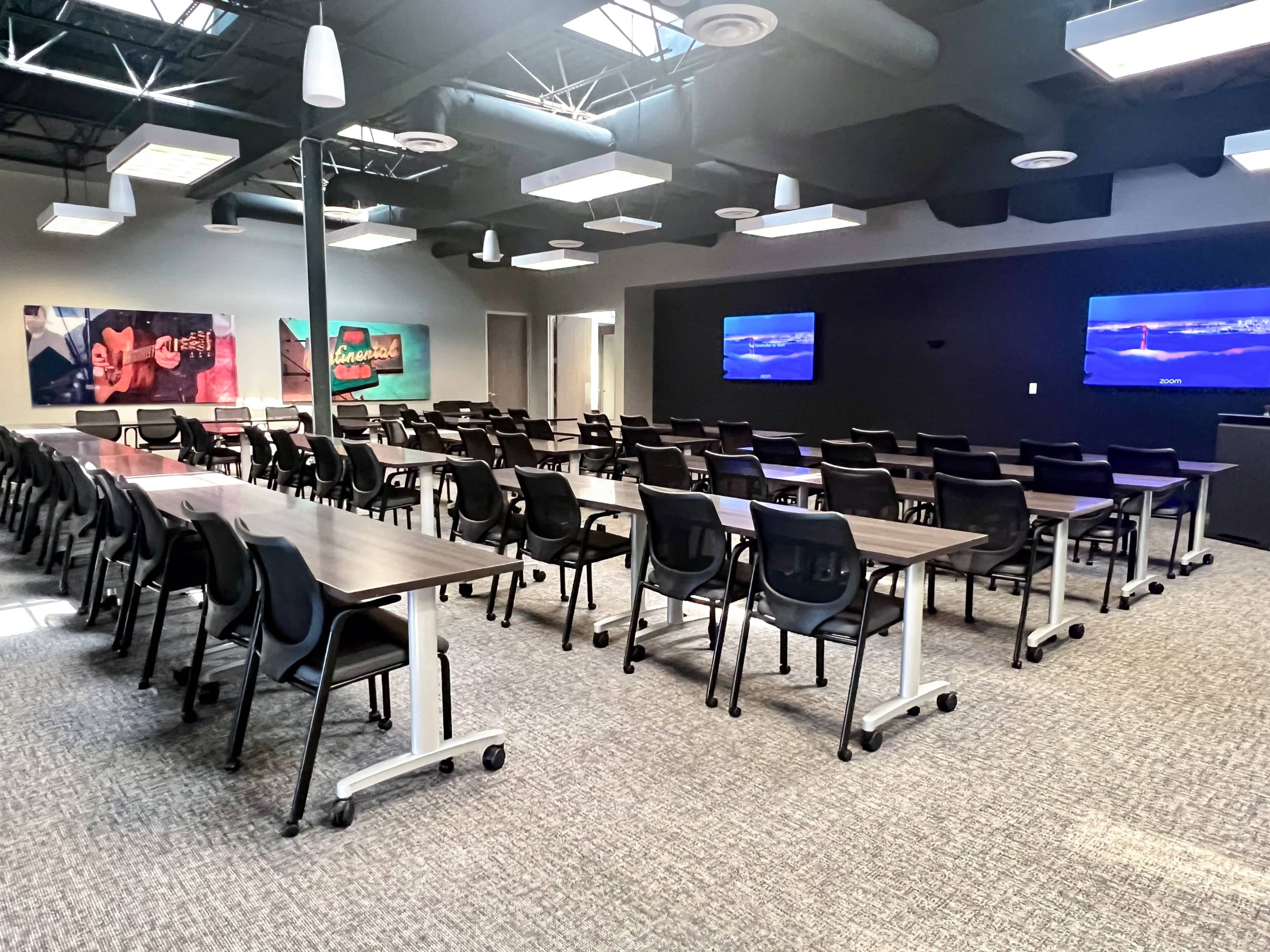 The image shows a spacious classroom set up with rows of tables and chairs, featuring two screens mounted on the walls.