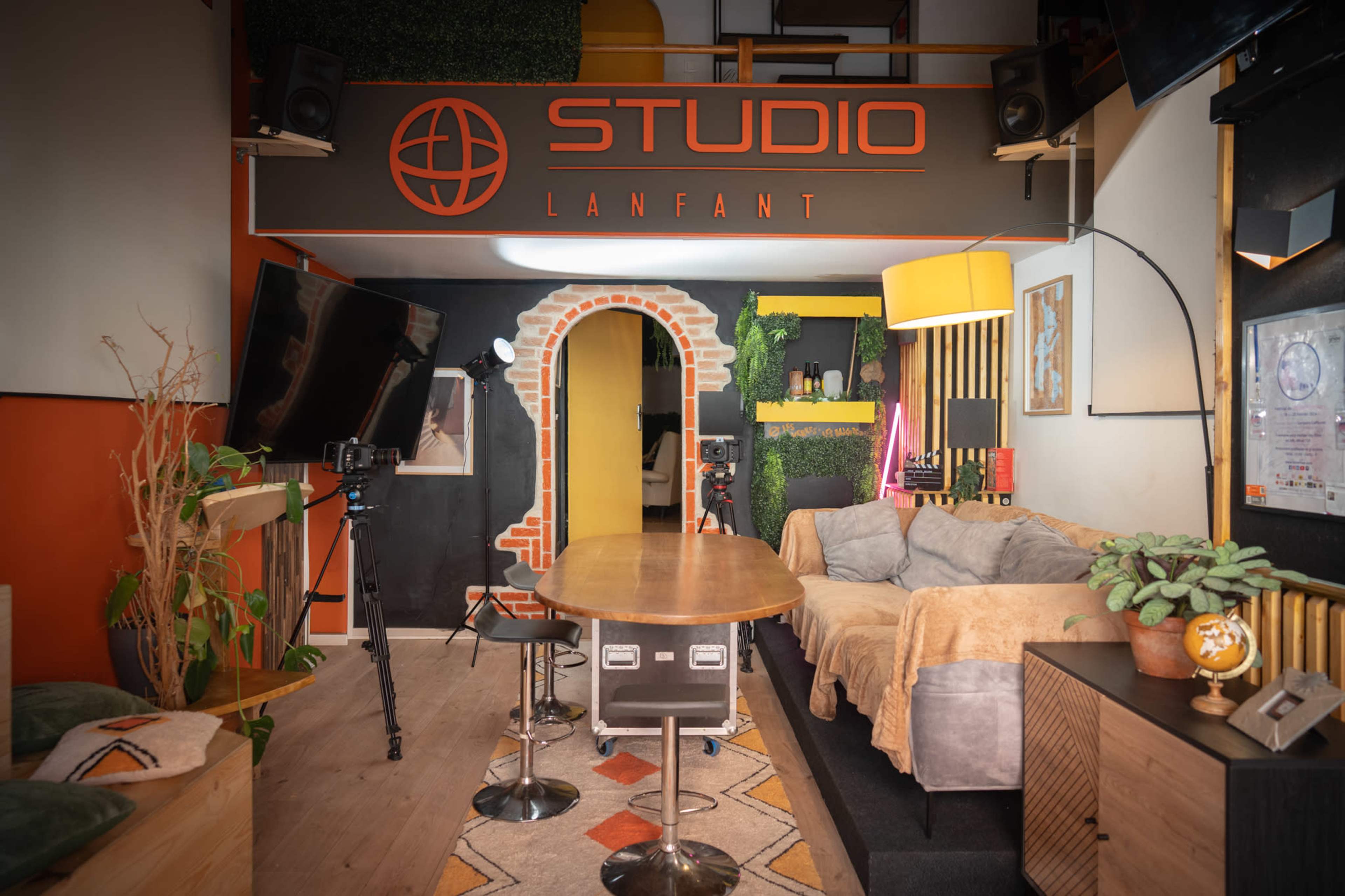 The image shows a studio space with a large table surrounded by chairs, a couch, decorative plants, and various media equipment, all under a sign that reads "STUDIO LANTFANT."