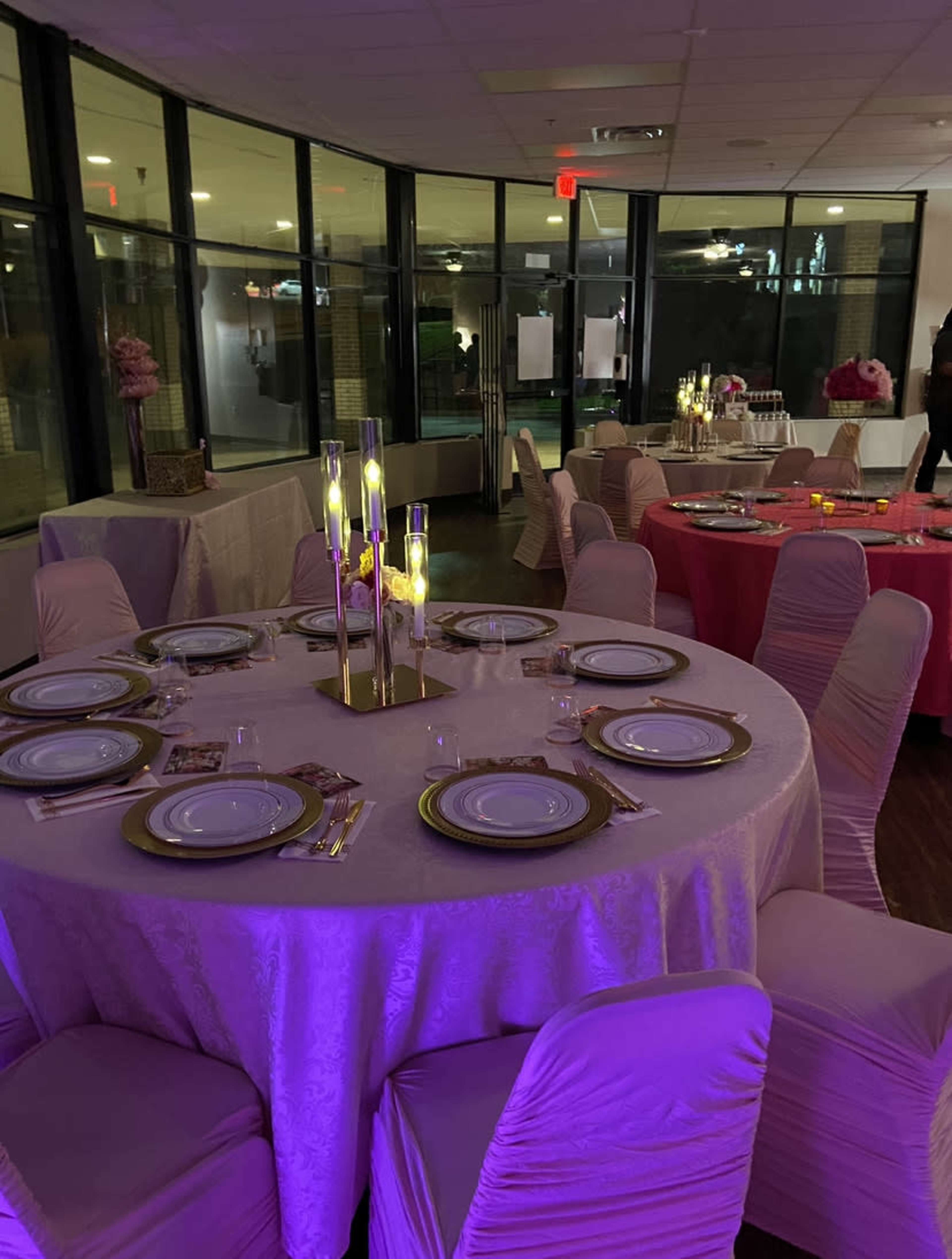 Elegantly set round tables with white plates and gold accents, surrounded by pink and light purple decor in a well-lit indoor venue.