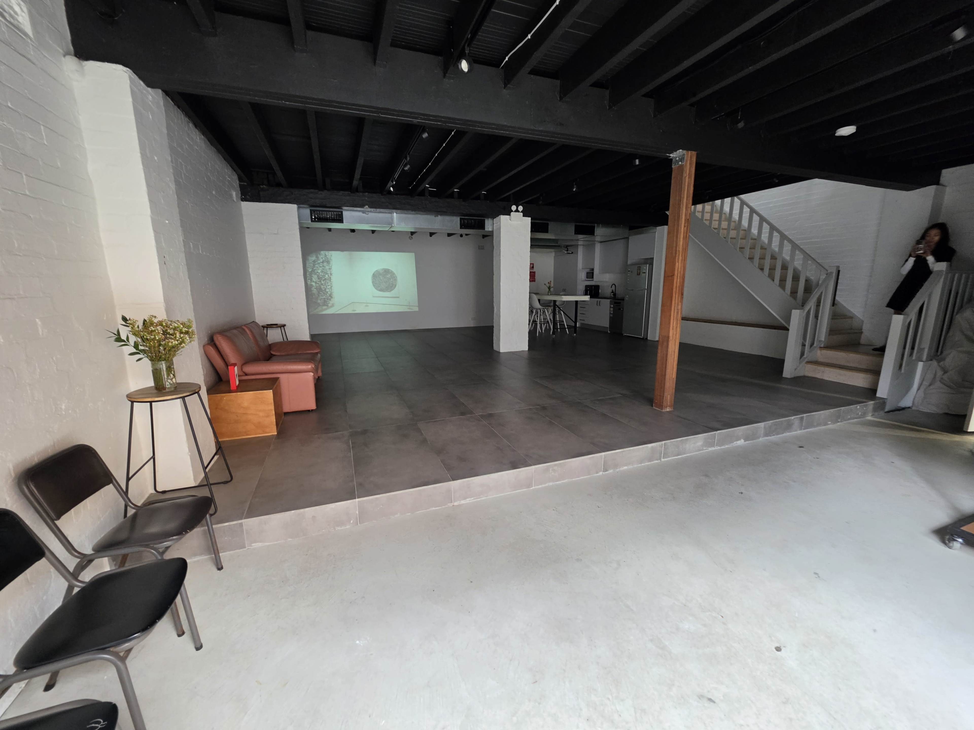 The image shows a spacious, empty room with a gray tiled floor, white brick walls, a projected image on a wall, and a staircase leading to an upper level.