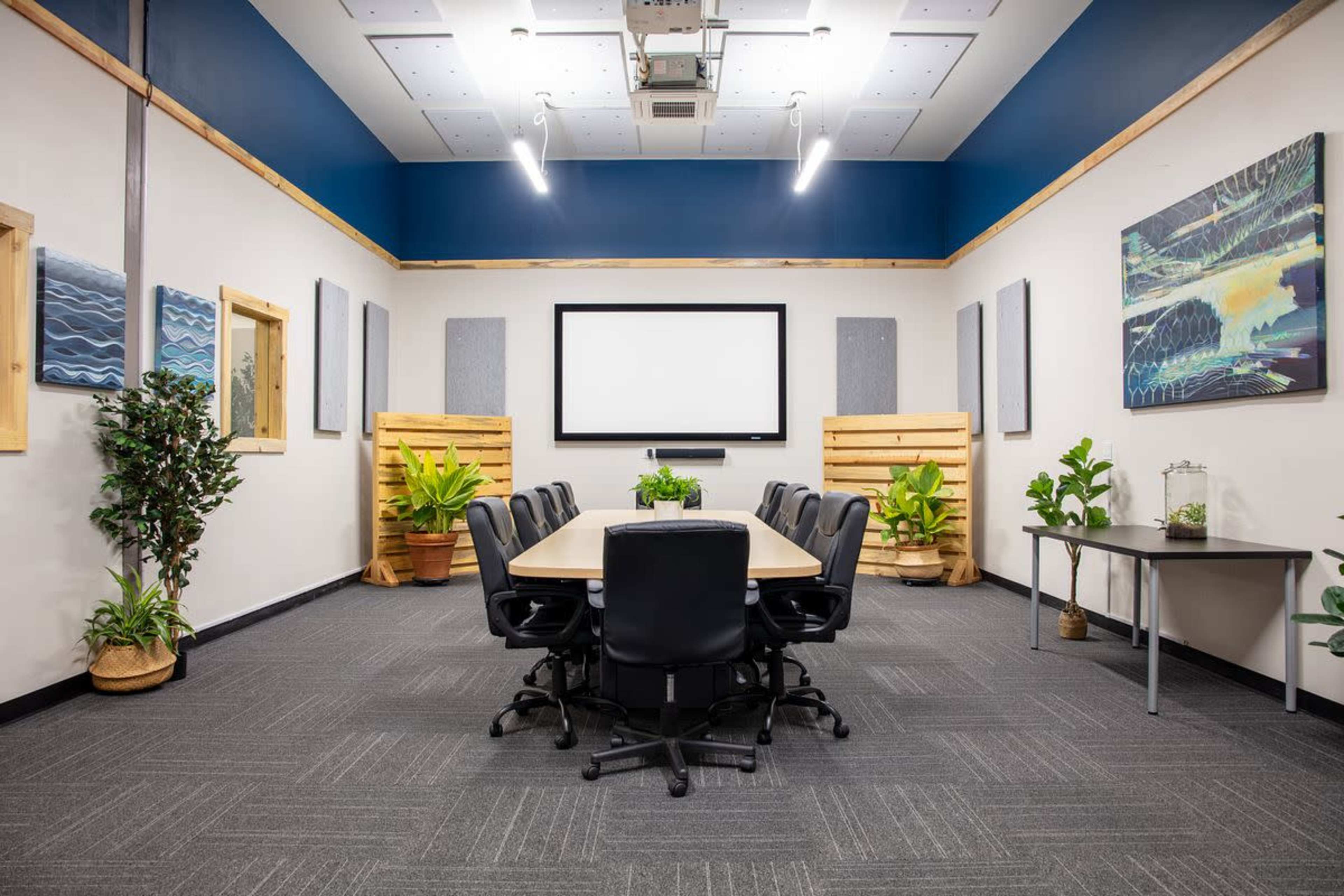 A conference room is equipped with a rectangular table and black chairs, flanked by plants and acoustic panels, with a large screen at the front and artwork on the walls.