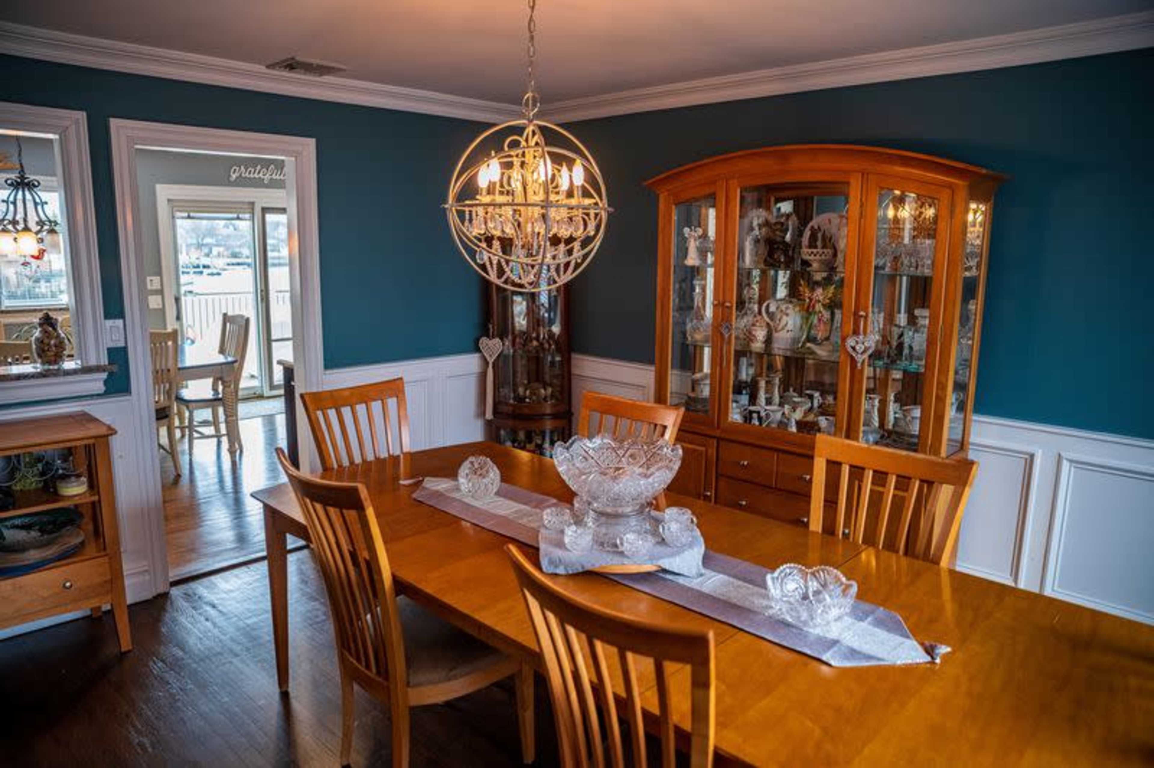 A wooden dining table with six chairs is set in a dining room featuring a china cabinet and a blue-painted wall.
