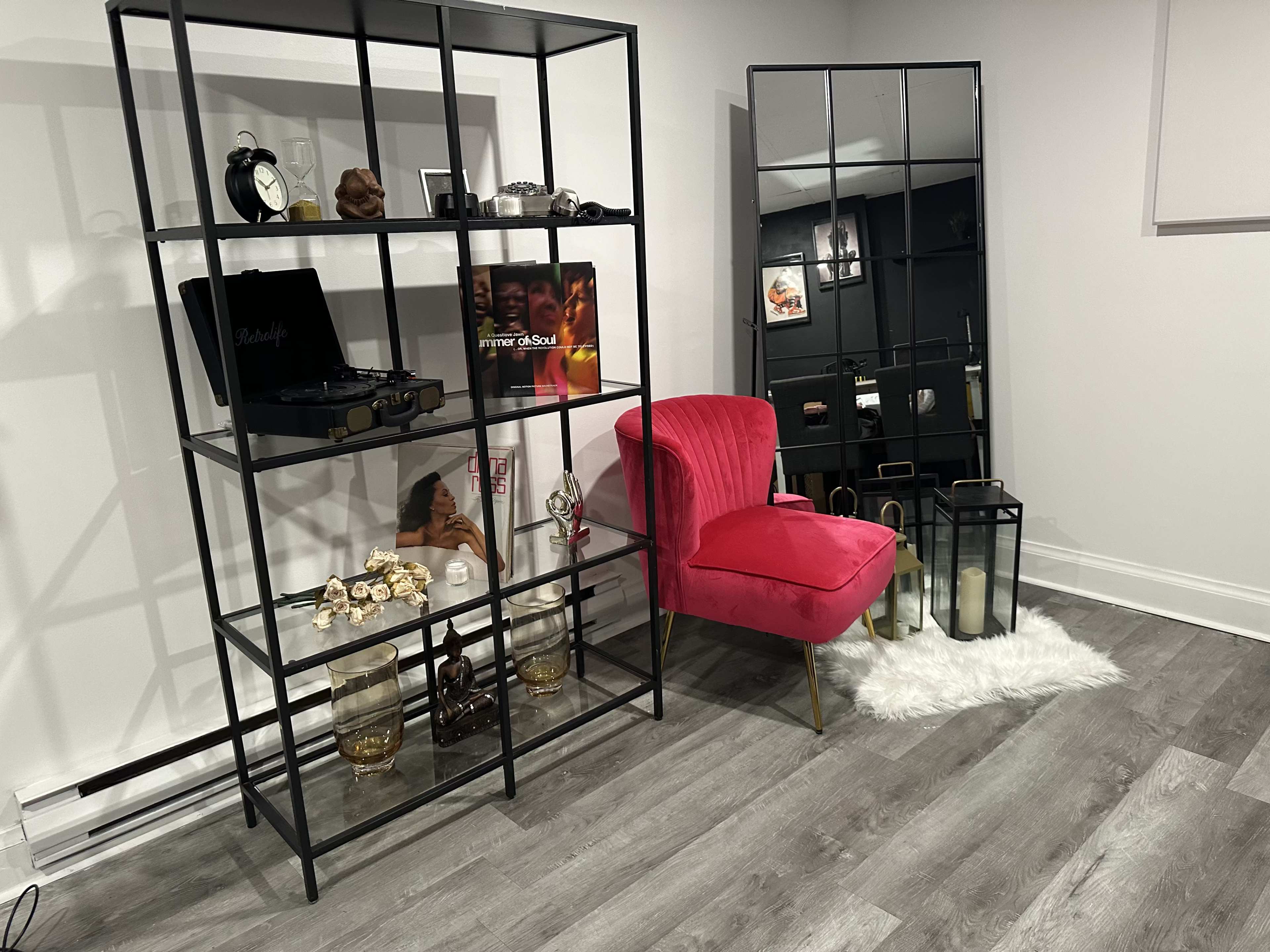 A modern interior space featuring a black metal shelving unit with decorative items, a pink velvet chair, a full-length mirror, and a soft rug on the floor.