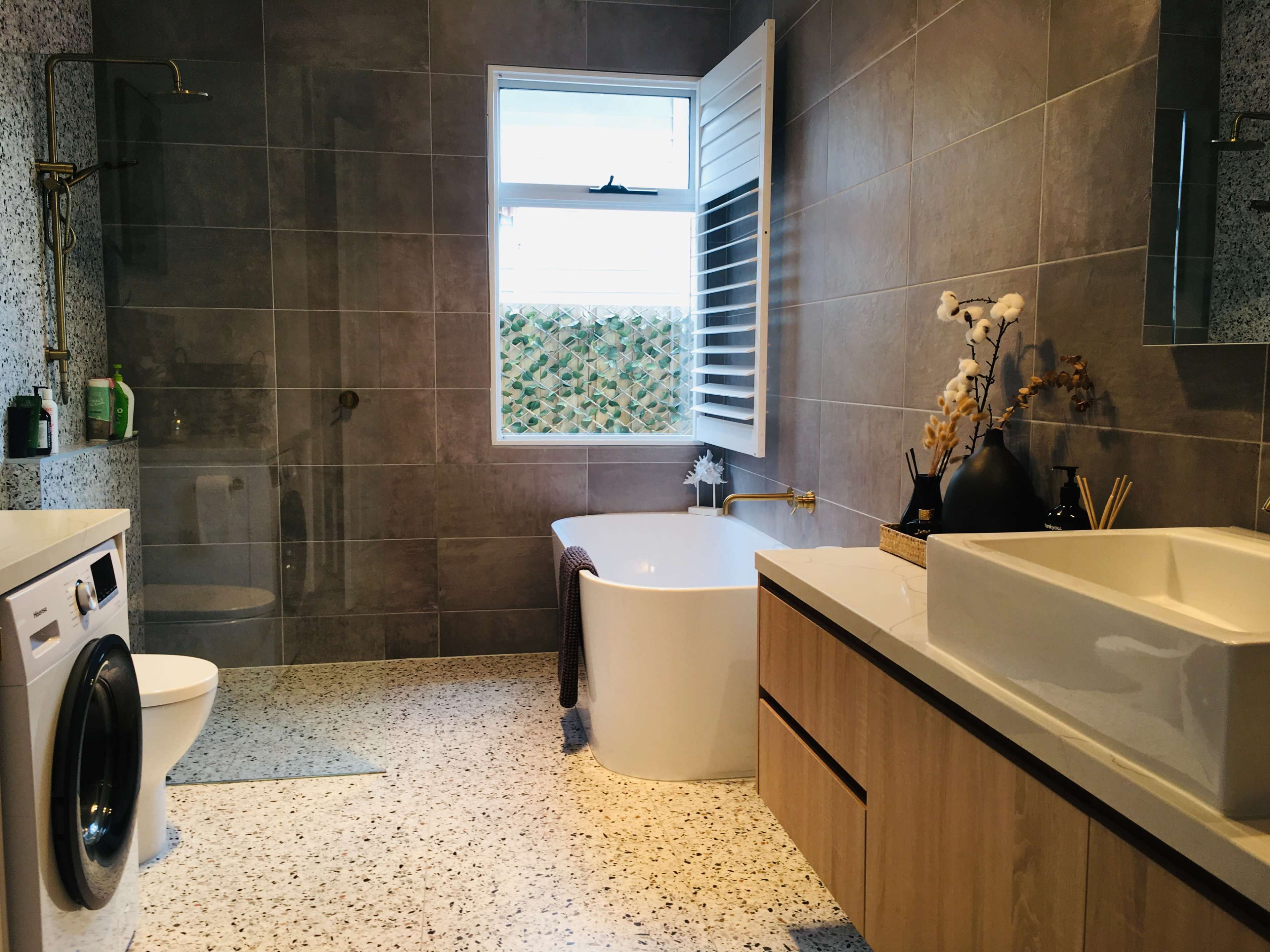 The modern bathroom features a freestanding bathtub, a washer/dryer unit, a sink with a wooden vanity, and a window with decorative shutters.