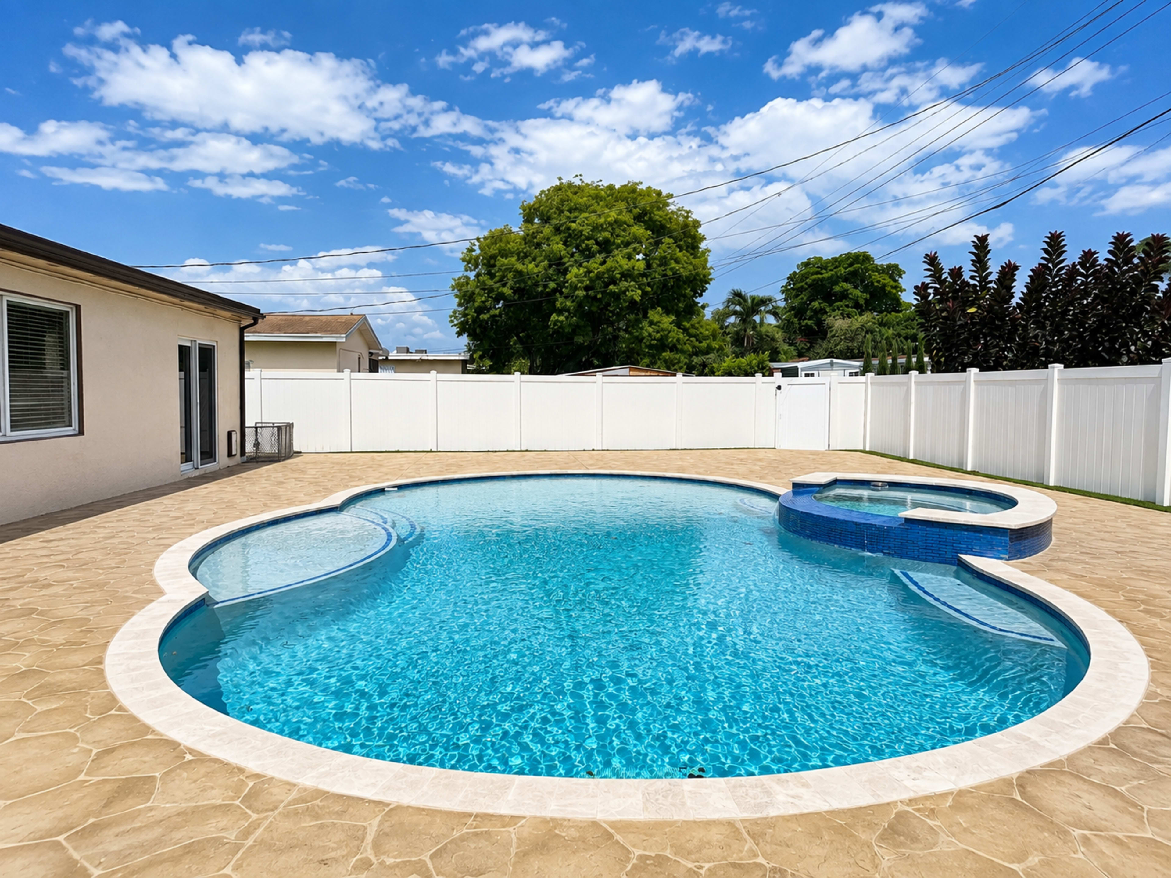The image shows a clear swimming pool with a attached spa area, surrounded by a stone deck and enclosed by a white fence under a blue sky.