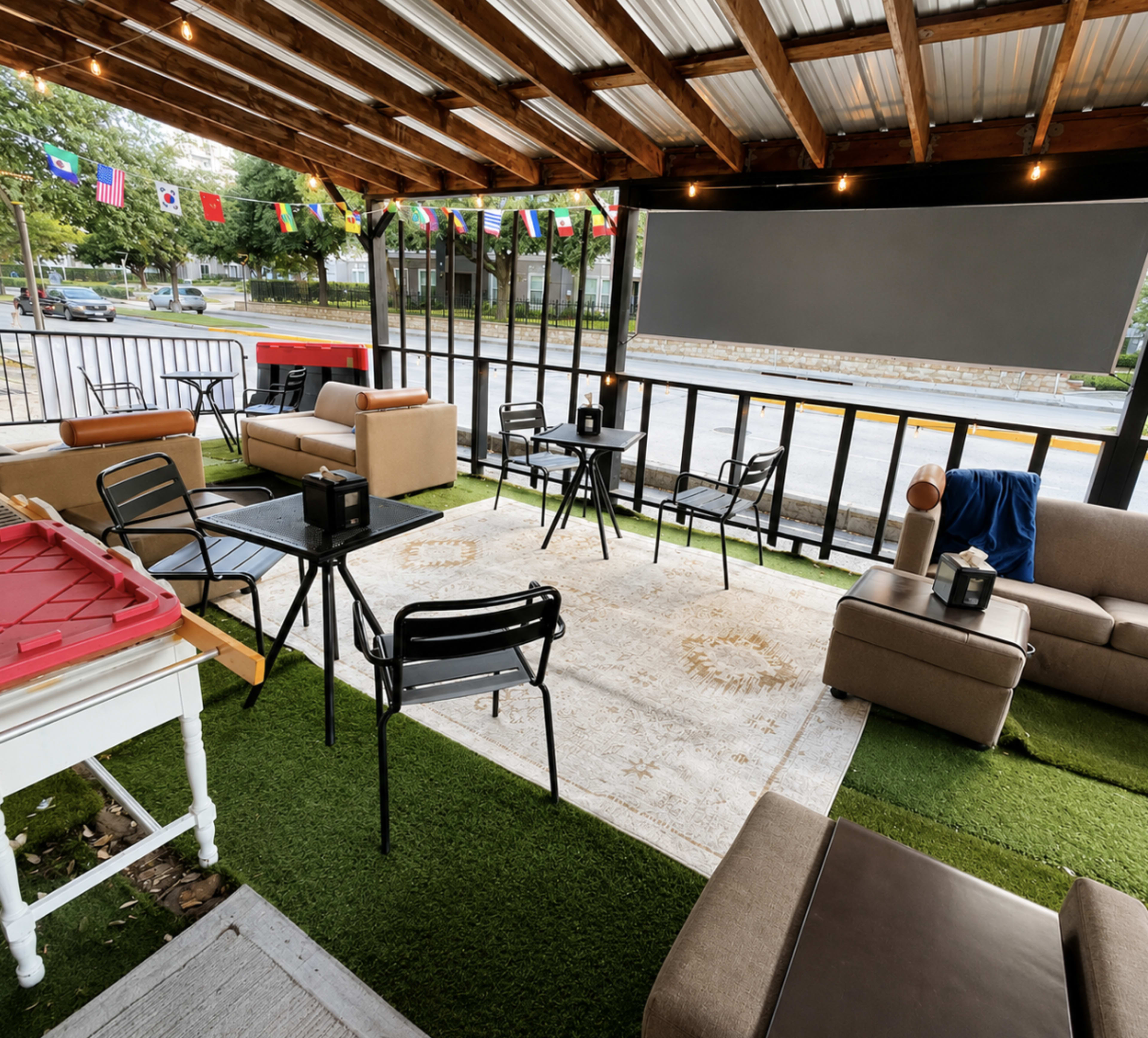 The image shows an outdoor seating area with multiple chairs and tables, a rug on artificial grass, and a large screen on a wooden structure, decorated with flags.