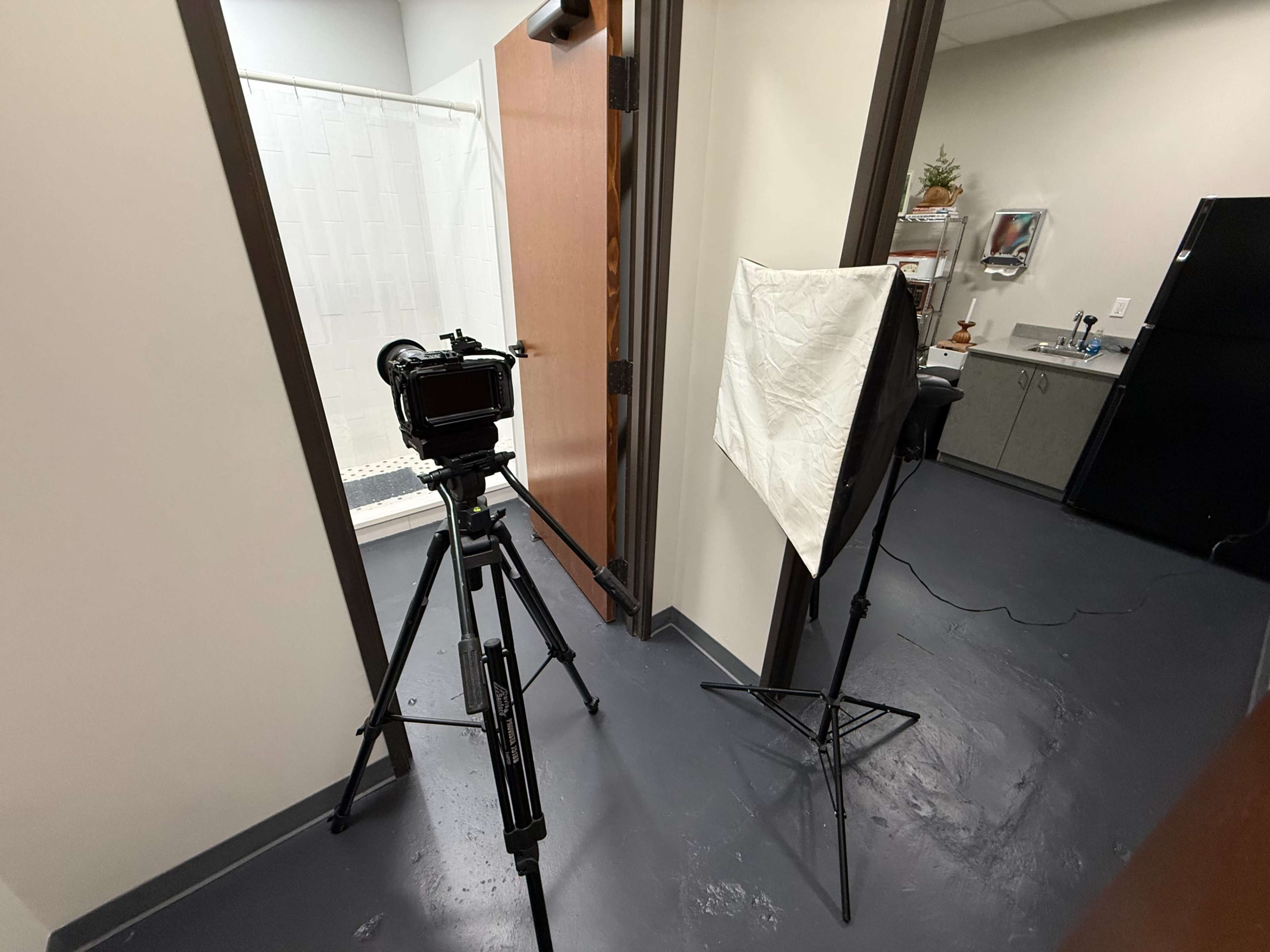 A camera on a tripod is set up in a hallway, facing an open door leading to a bathroom with a shower curtain, while a softbox light is positioned nearby.