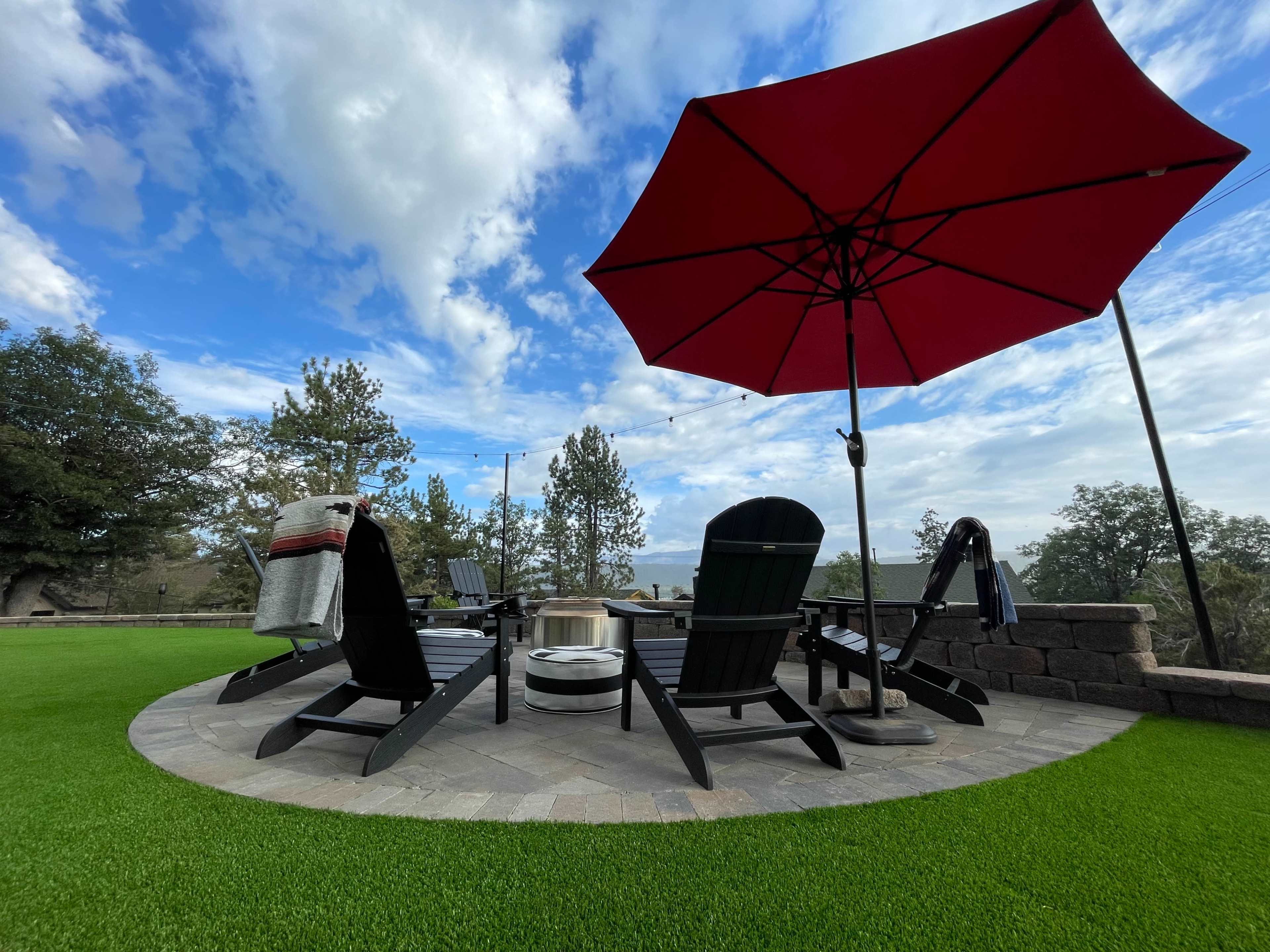 A circular stone patio features black Adirondack chairs, a fire pit, and a red umbrella on a lawn of artificial grass.