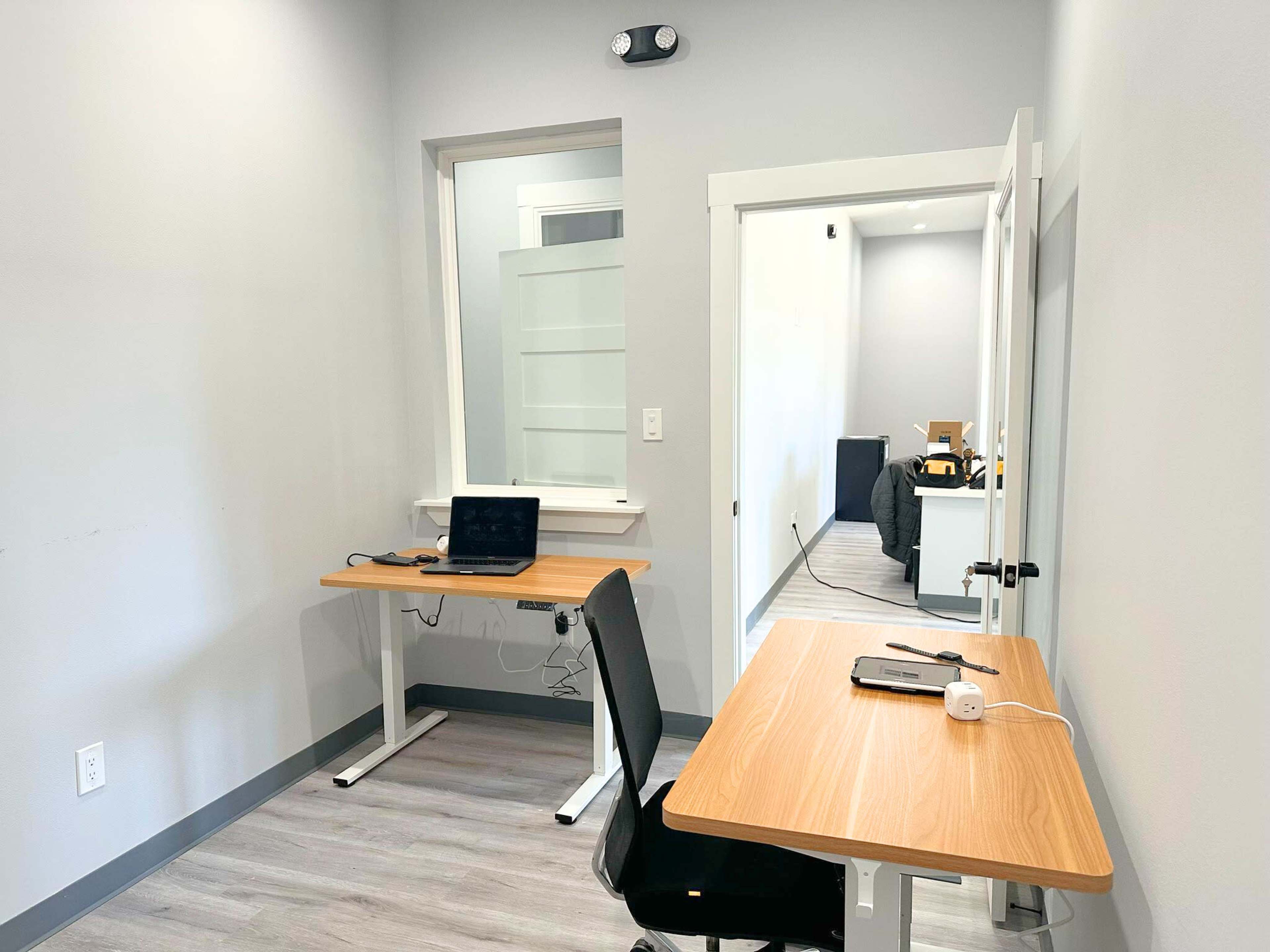 The image shows a small office space with two wooden desks, one equipped with a laptop, and a doorway leading to another room in the background.