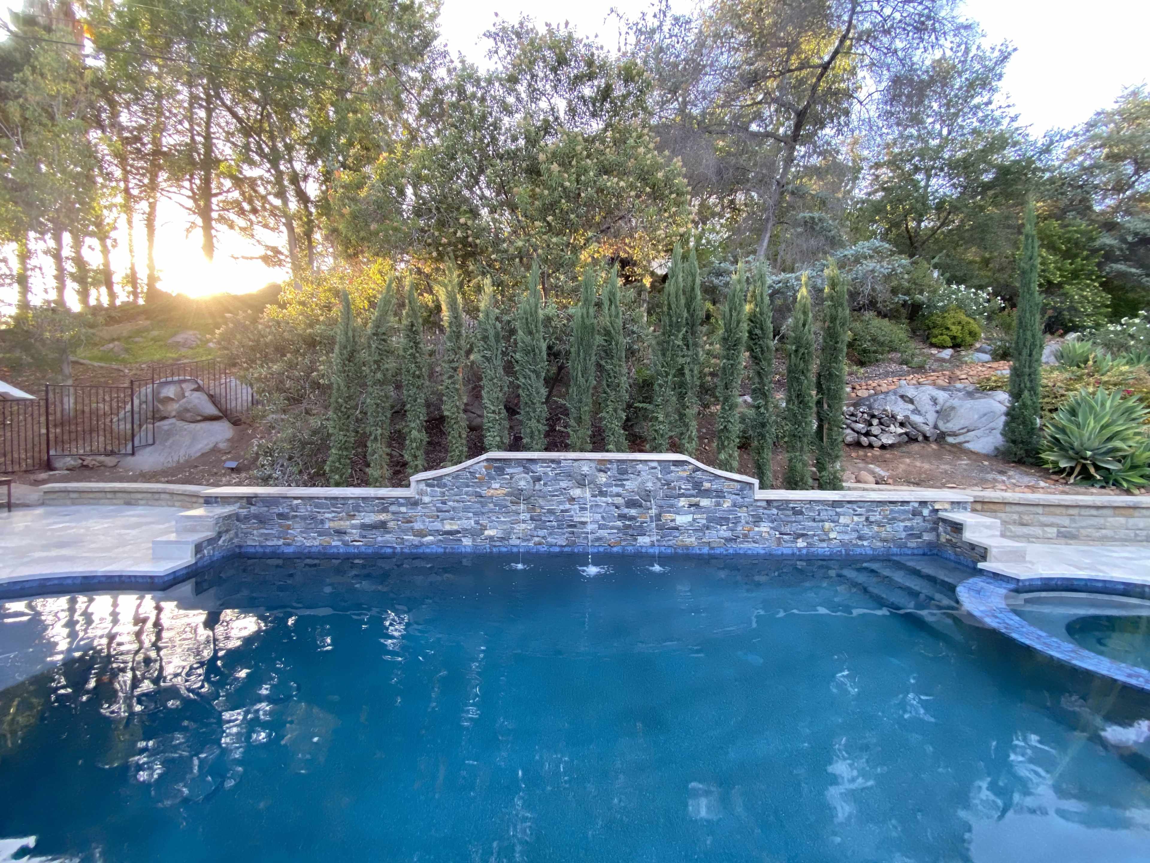 A backyard pool surrounded by tall greenery and stone landscaping, with a sunset visible in the background.