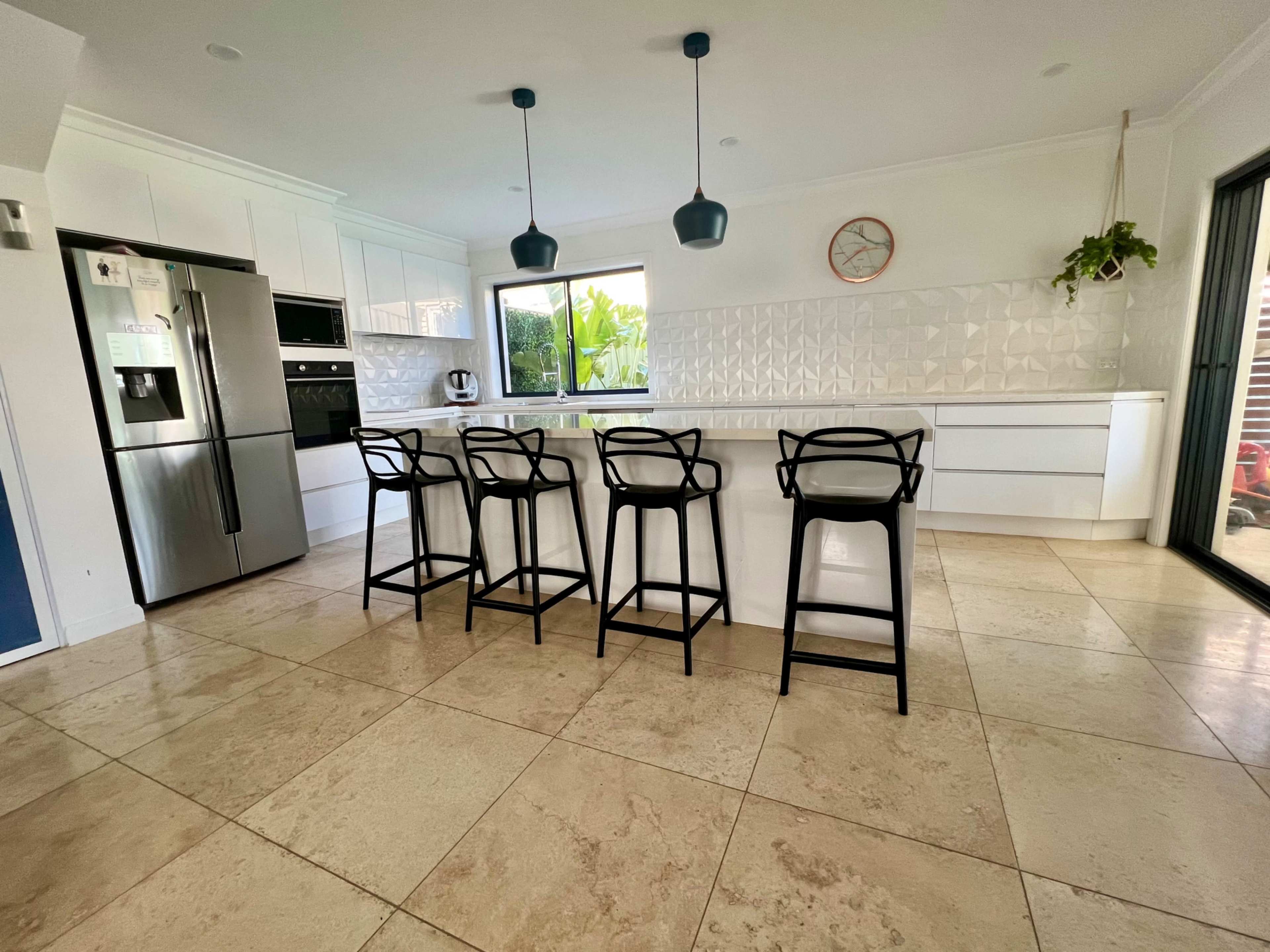 The image shows a modern kitchen with a large island and four black bar stools, featuring white cabinetry and a tiled backsplash.