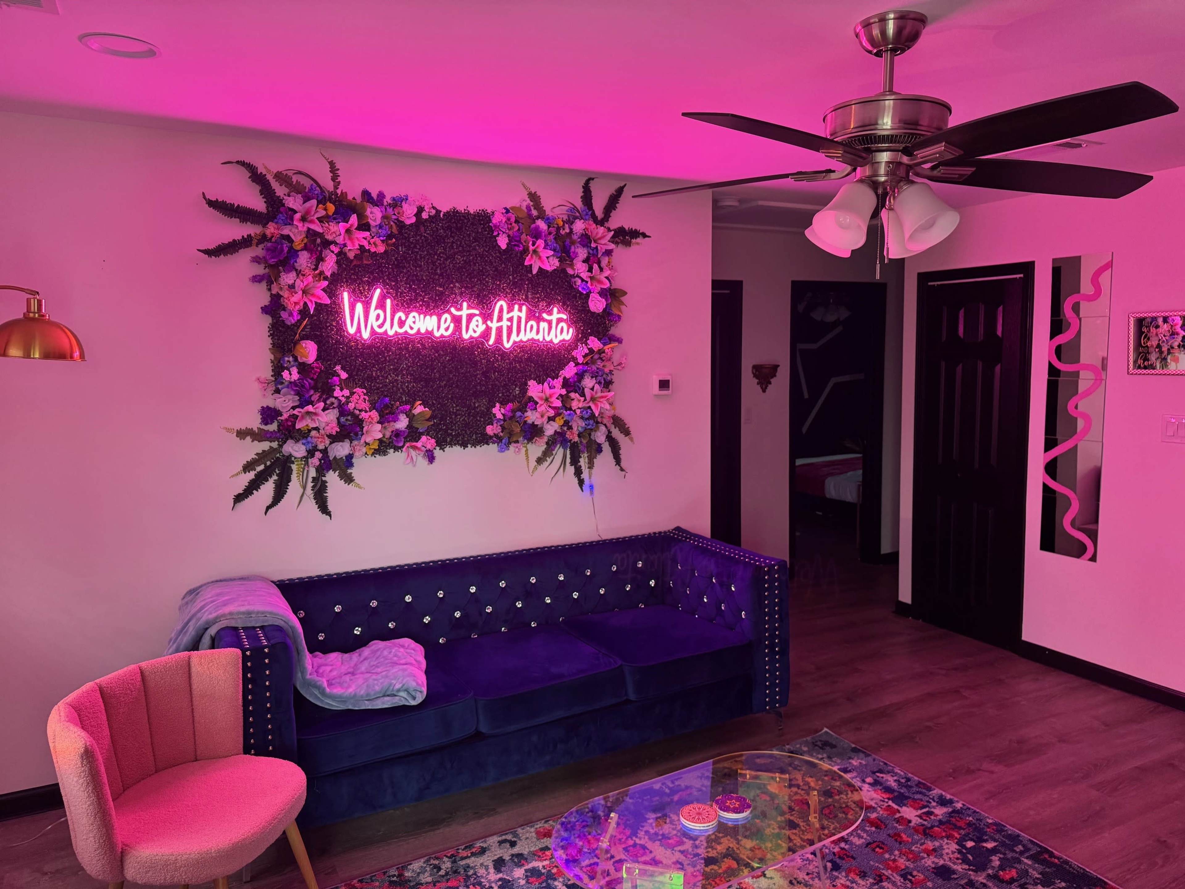A cozy interior space with a blue velvet couch, a pink neon sign that says "Welcome to Atlanta," and colorful decor elements including a patterned rug and modern lighting.
