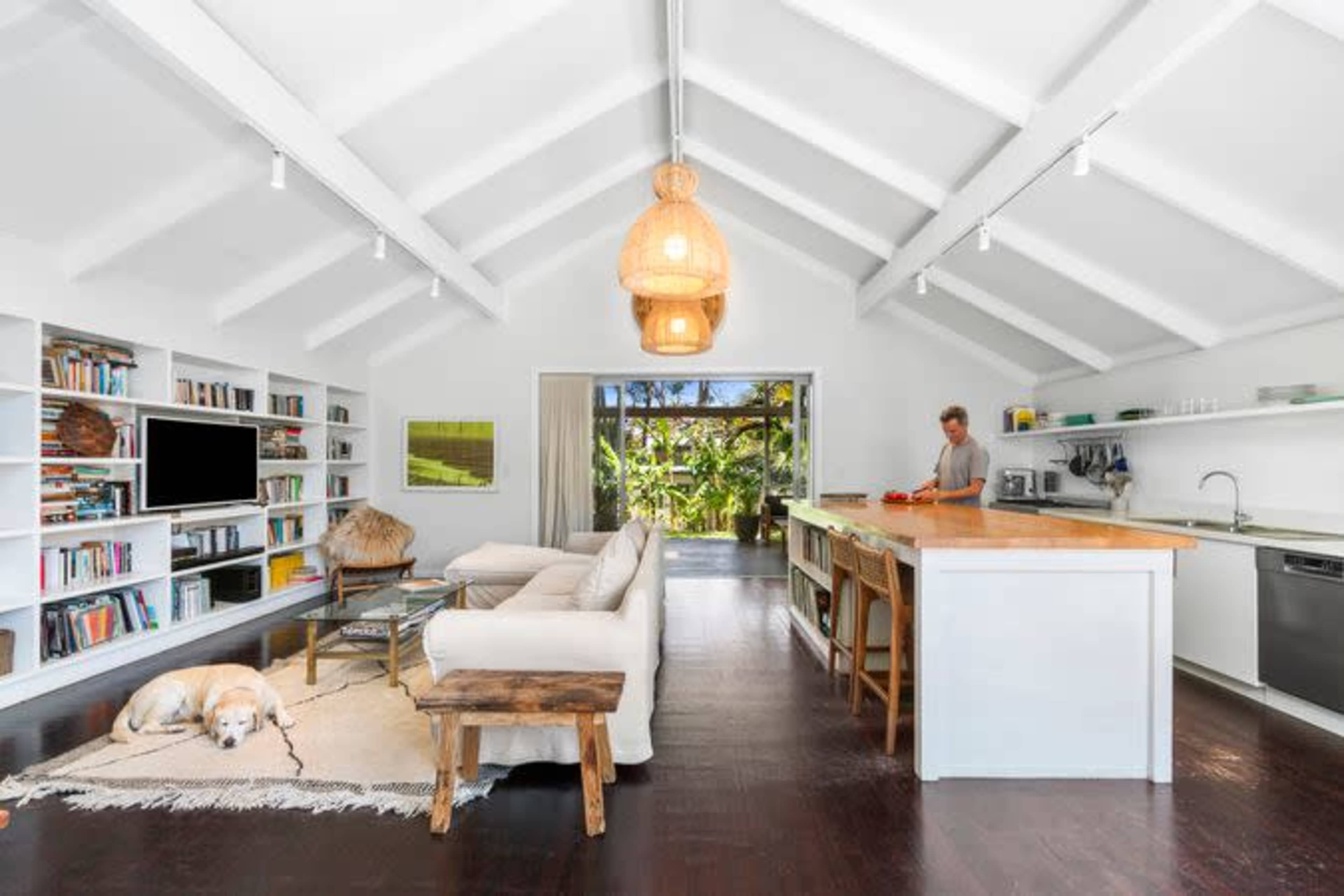 A modern living room features a white couch, a wooden coffee table, shelves filled with books, and a kitchen area with a counter where a person is preparing food.