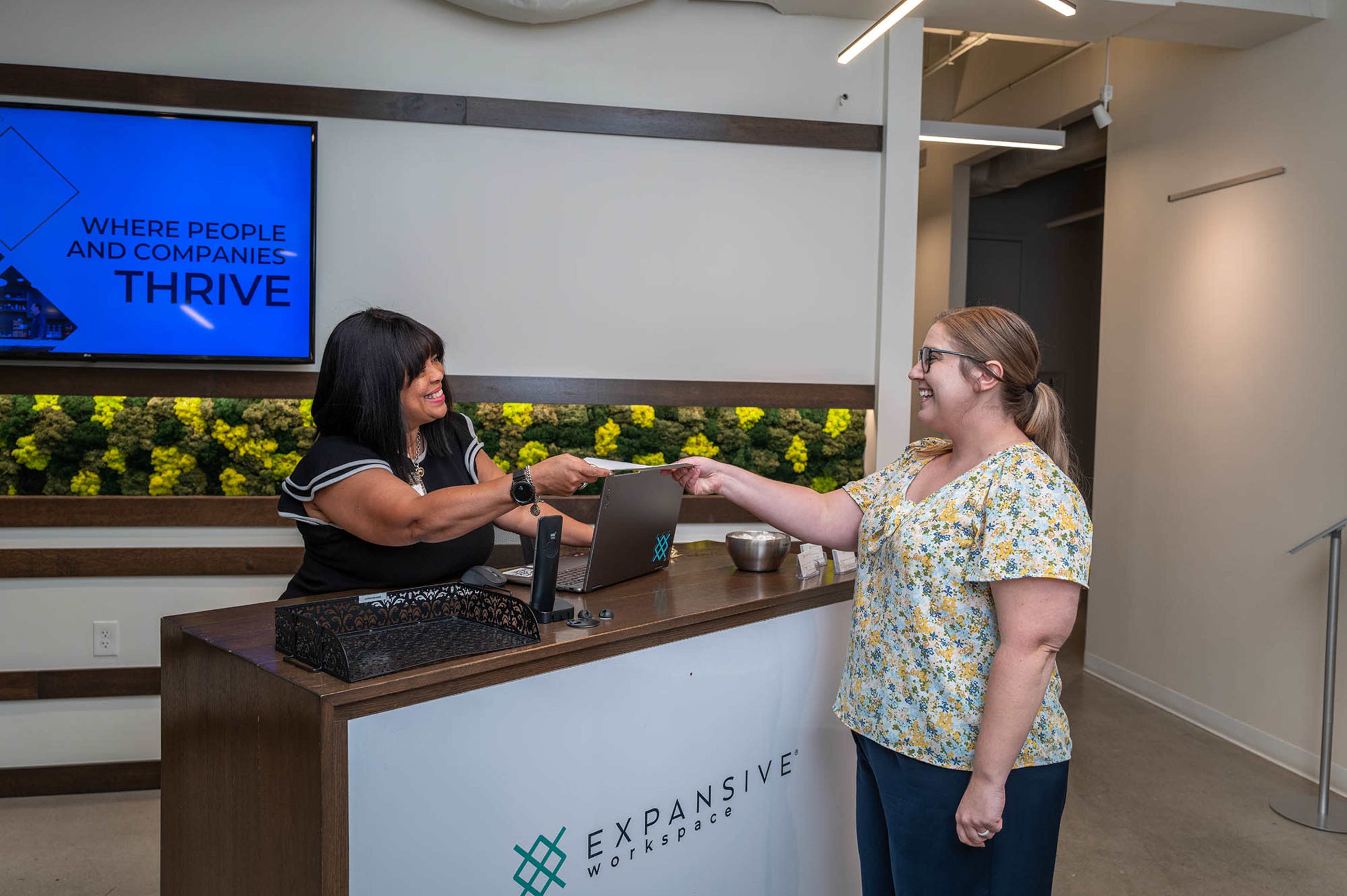 A receptionist hands a visitor a keycard at a modern co-working space reception desk.