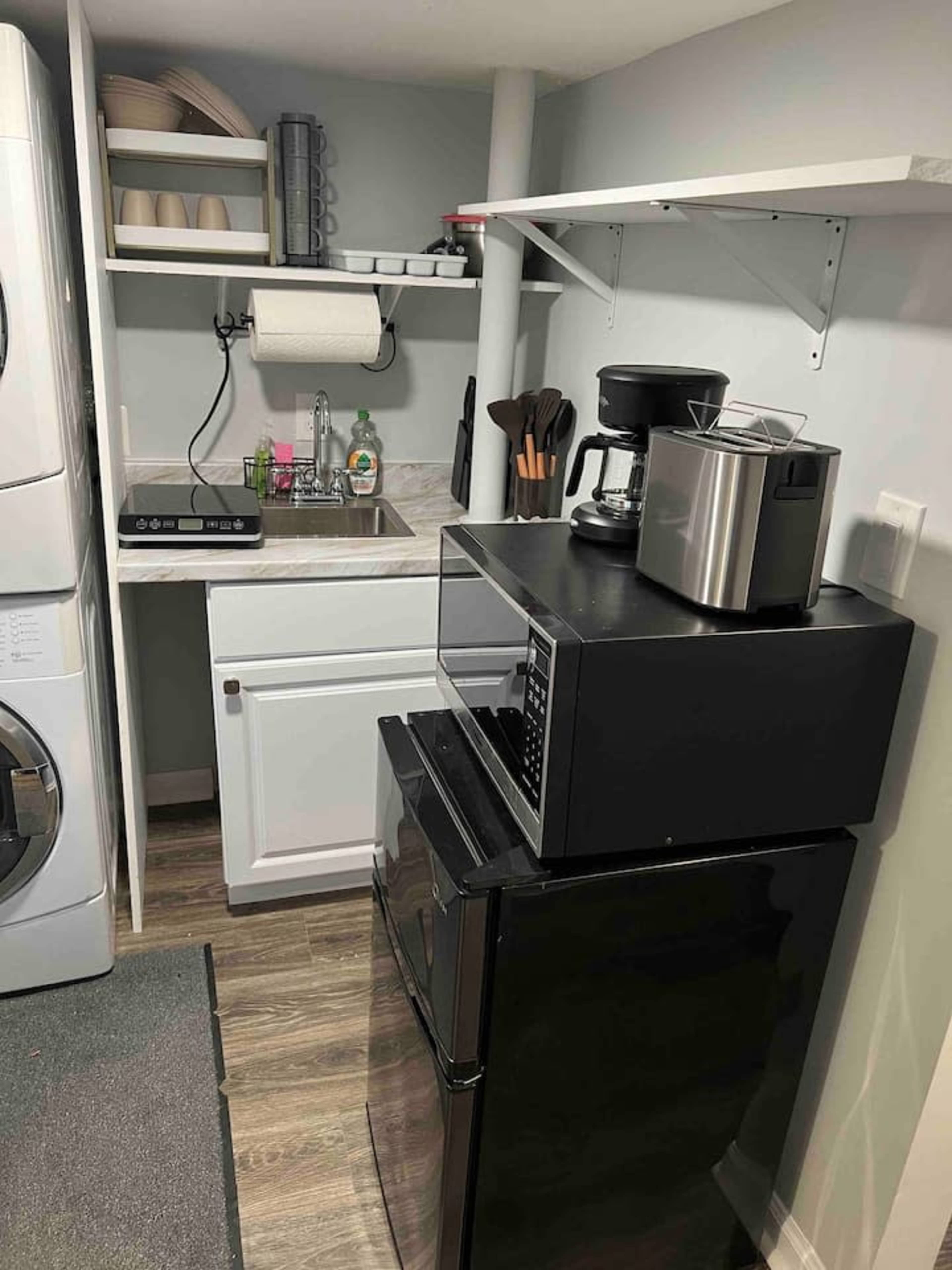 A compact kitchen area featuring a refrigerator, microwave, coffee maker, and a small sink with dishware above.