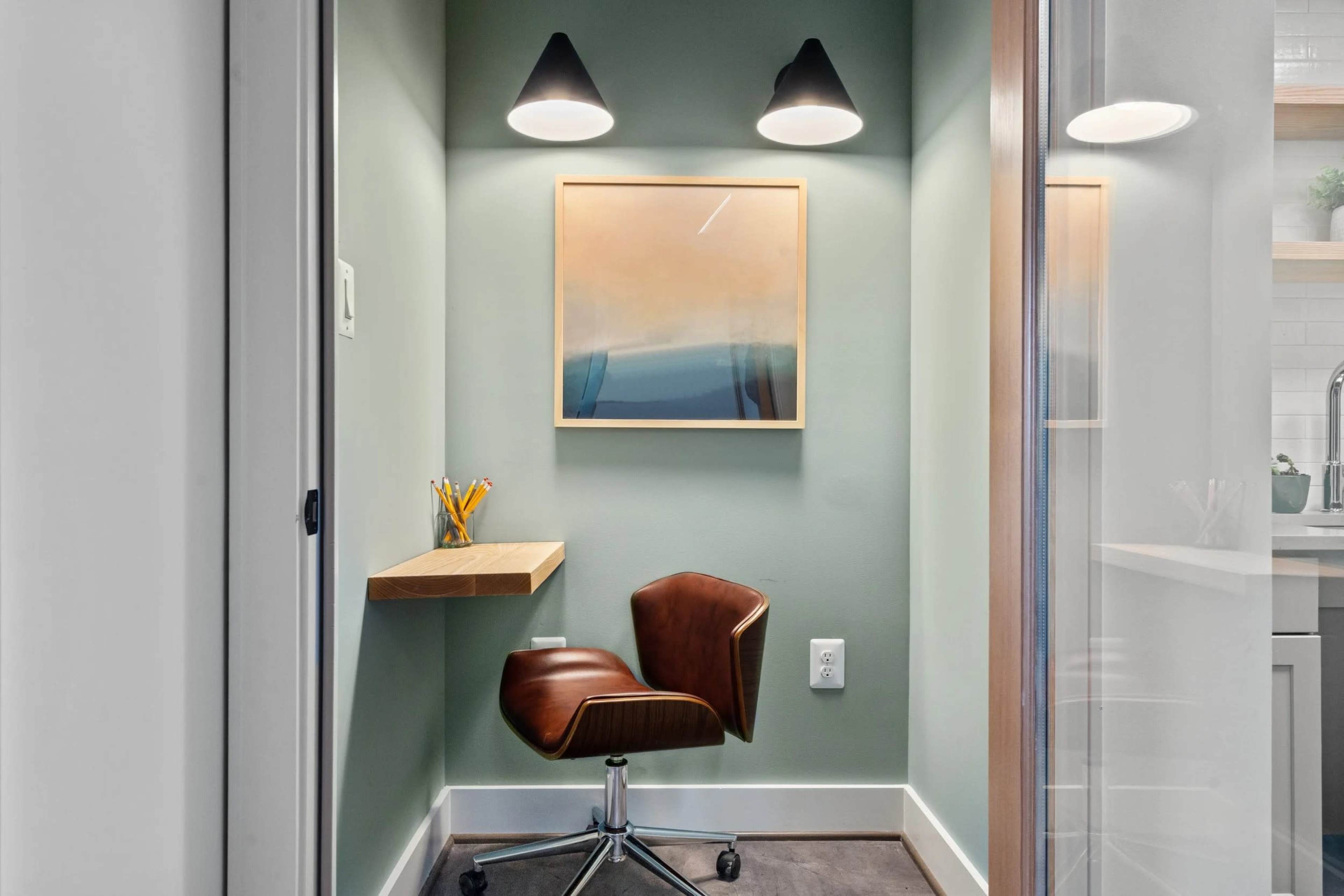 The image shows a small office nook with a wooden desk, an ergonomic chair, and a framed abstract painting mounted on a green wall, illuminated by two wall-mounted lights.