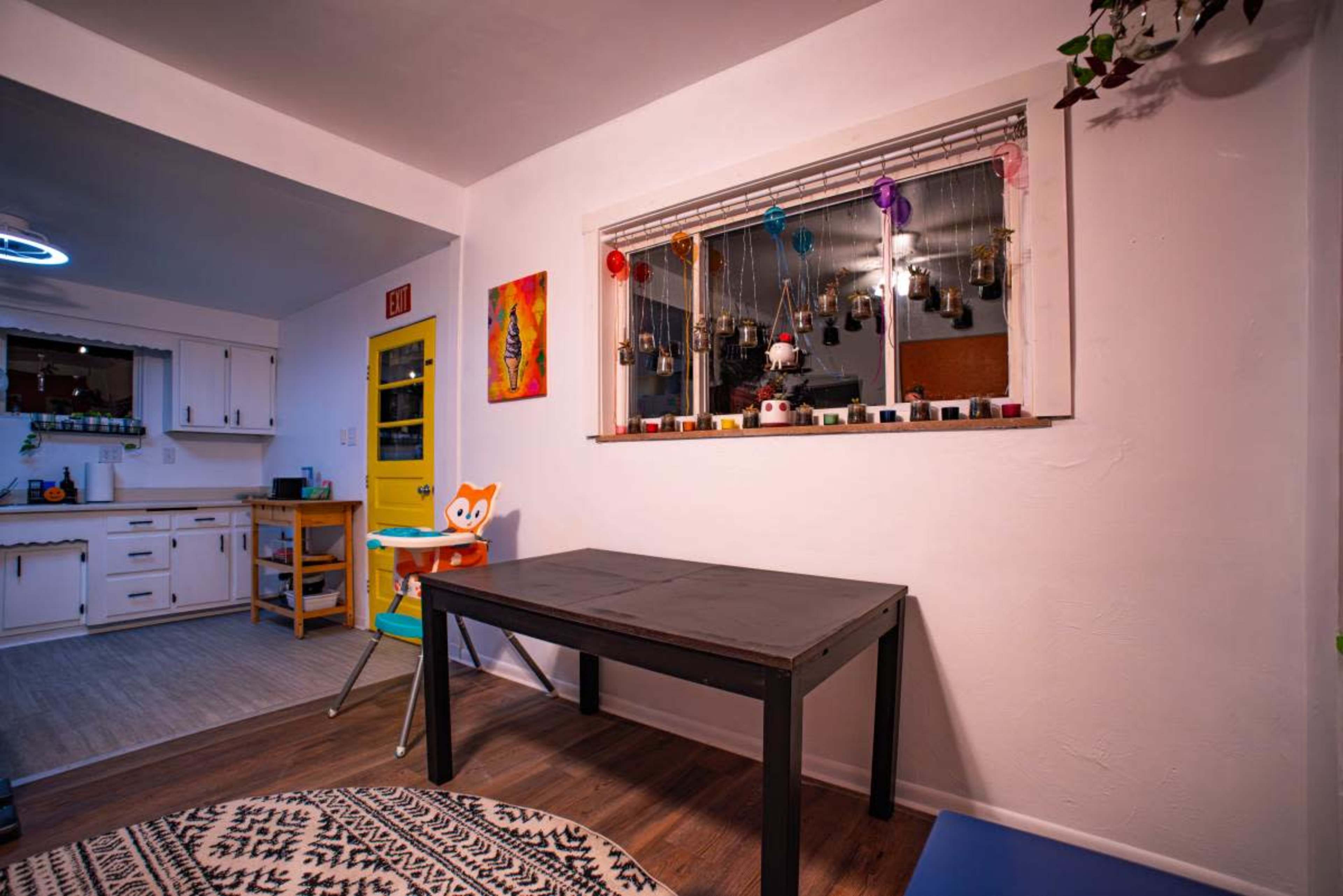 The image shows a corner of a room featuring a black table, a window adorned with hanging decorations, and a glimpse of a kitchen area in the background.