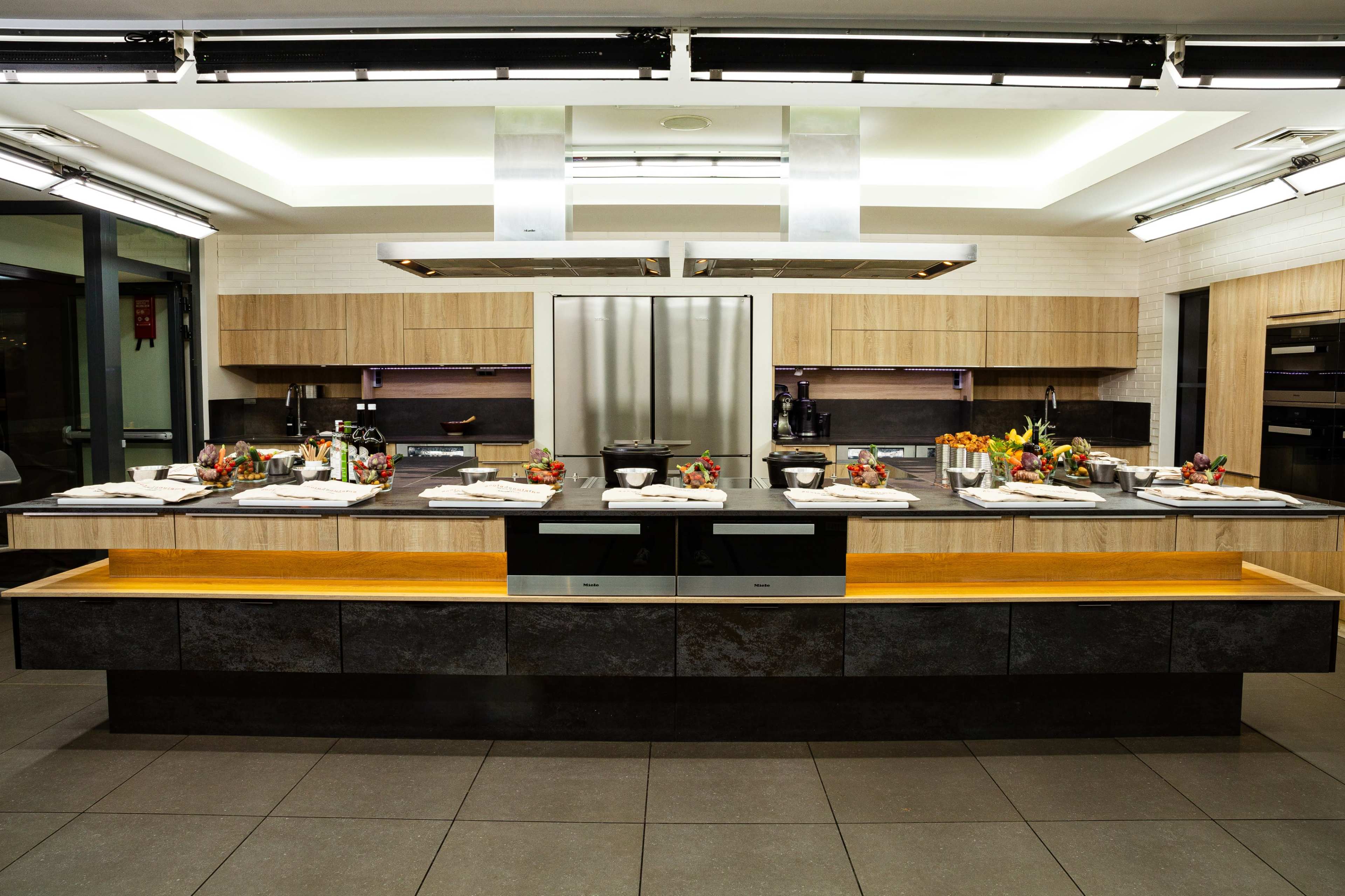 The image shows a modern kitchen with a large central island, sleek cabinetry, and various cooking appliances arranged in a spacious layout.