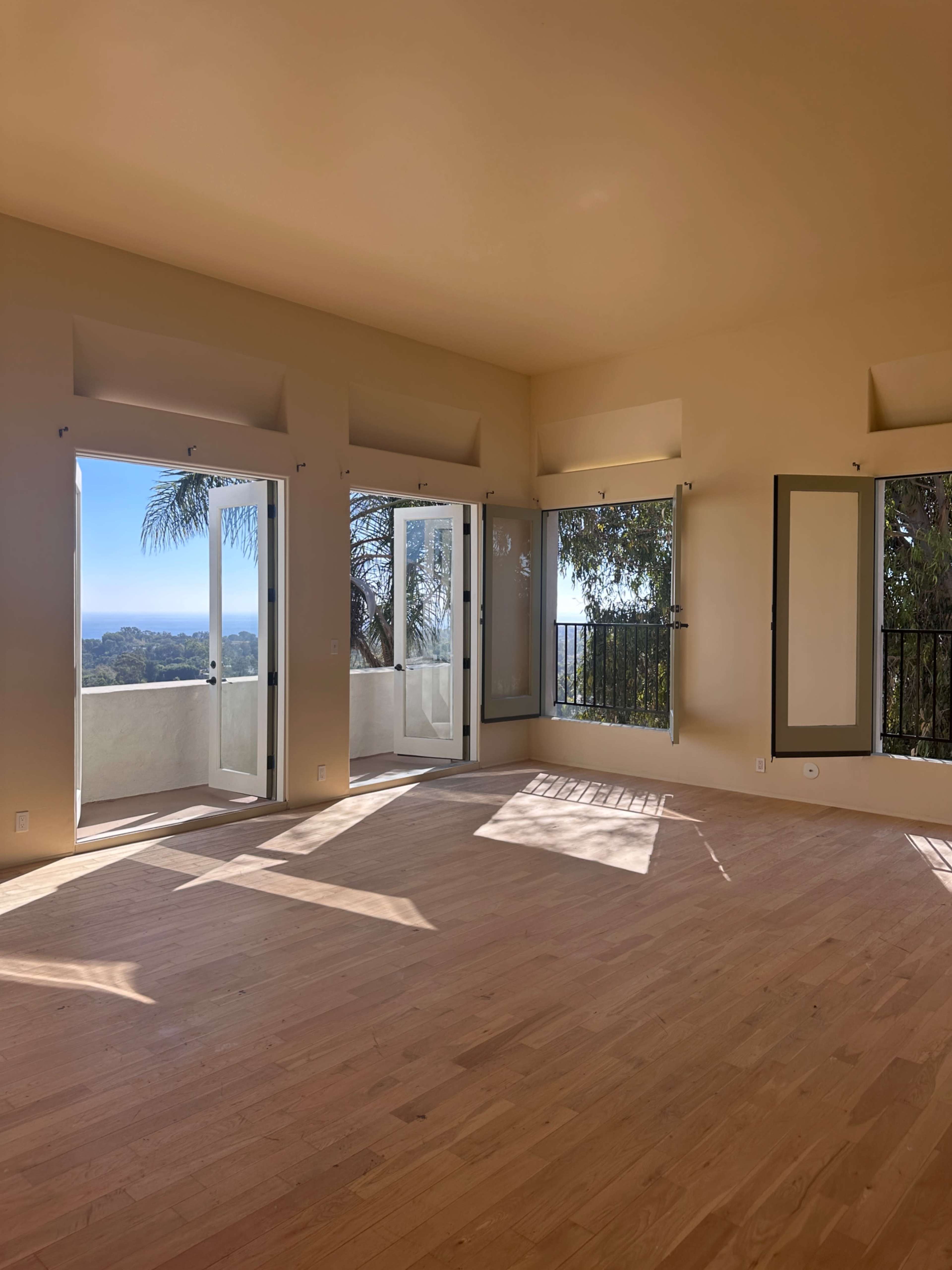 The image shows a spacious interior room with light wooden flooring, large windows, and double doors that open to a scenic view.