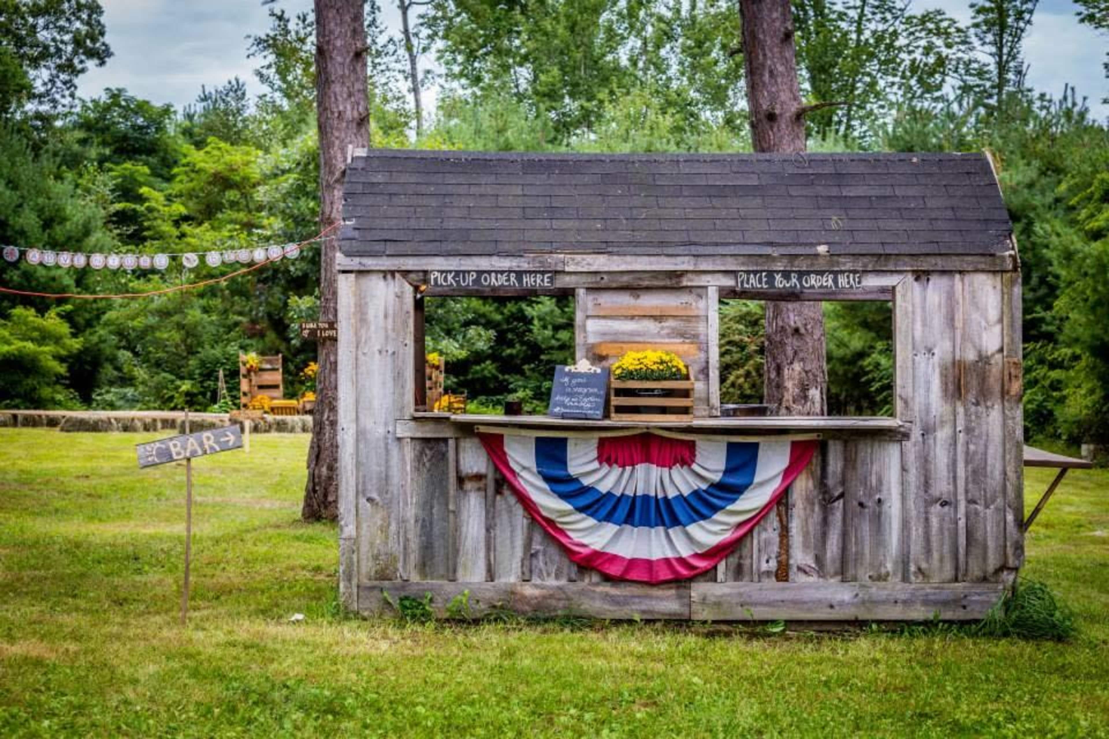The image shows a wooden kiosk decorated with red, white, and blue bunting, set in a grassy area surrounded by trees.
