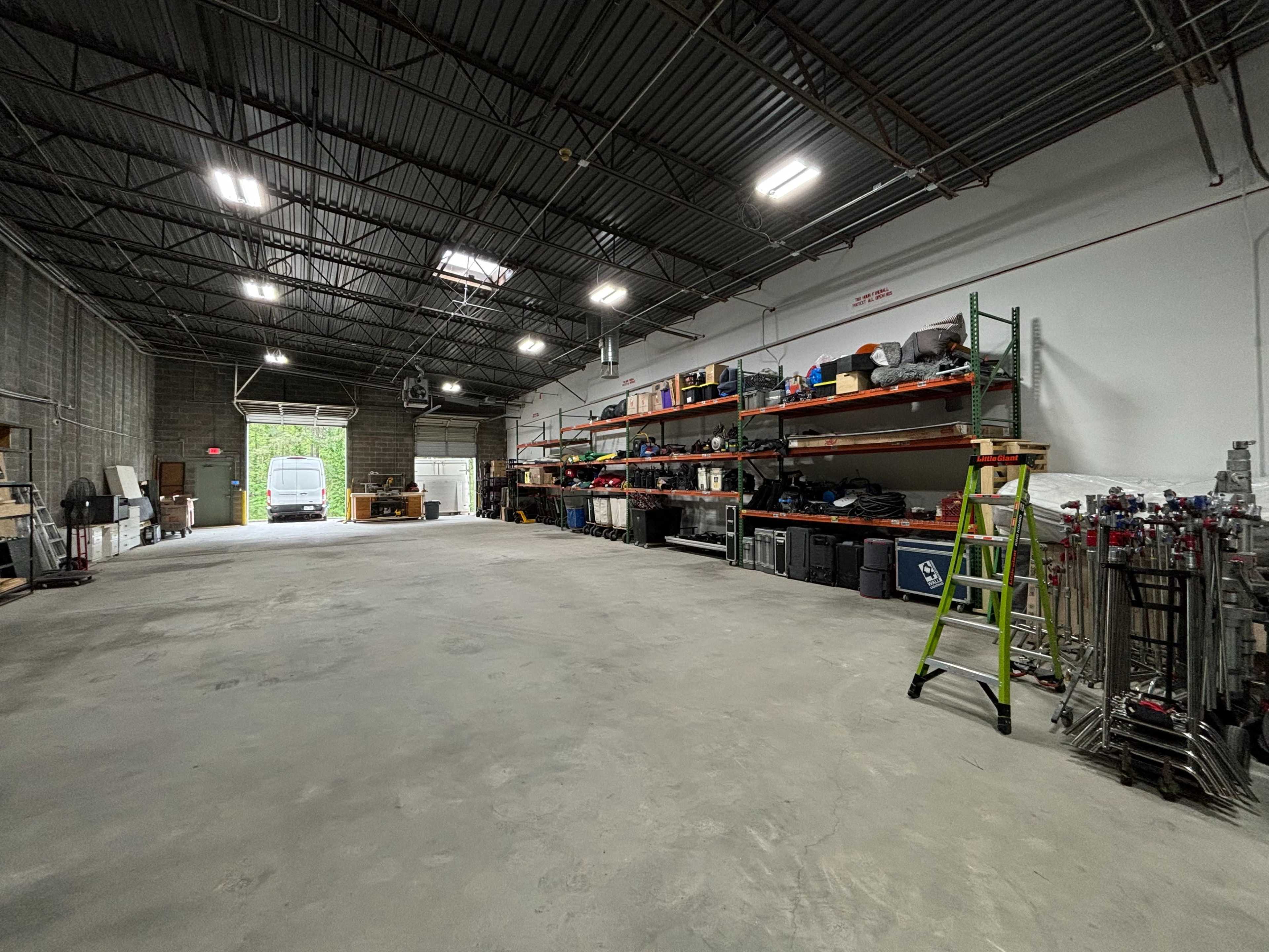 A spacious garage or warehouse interior with high ceilings, neatly arranged shelving units filled with various equipment, and a ladder positioned on the right side.