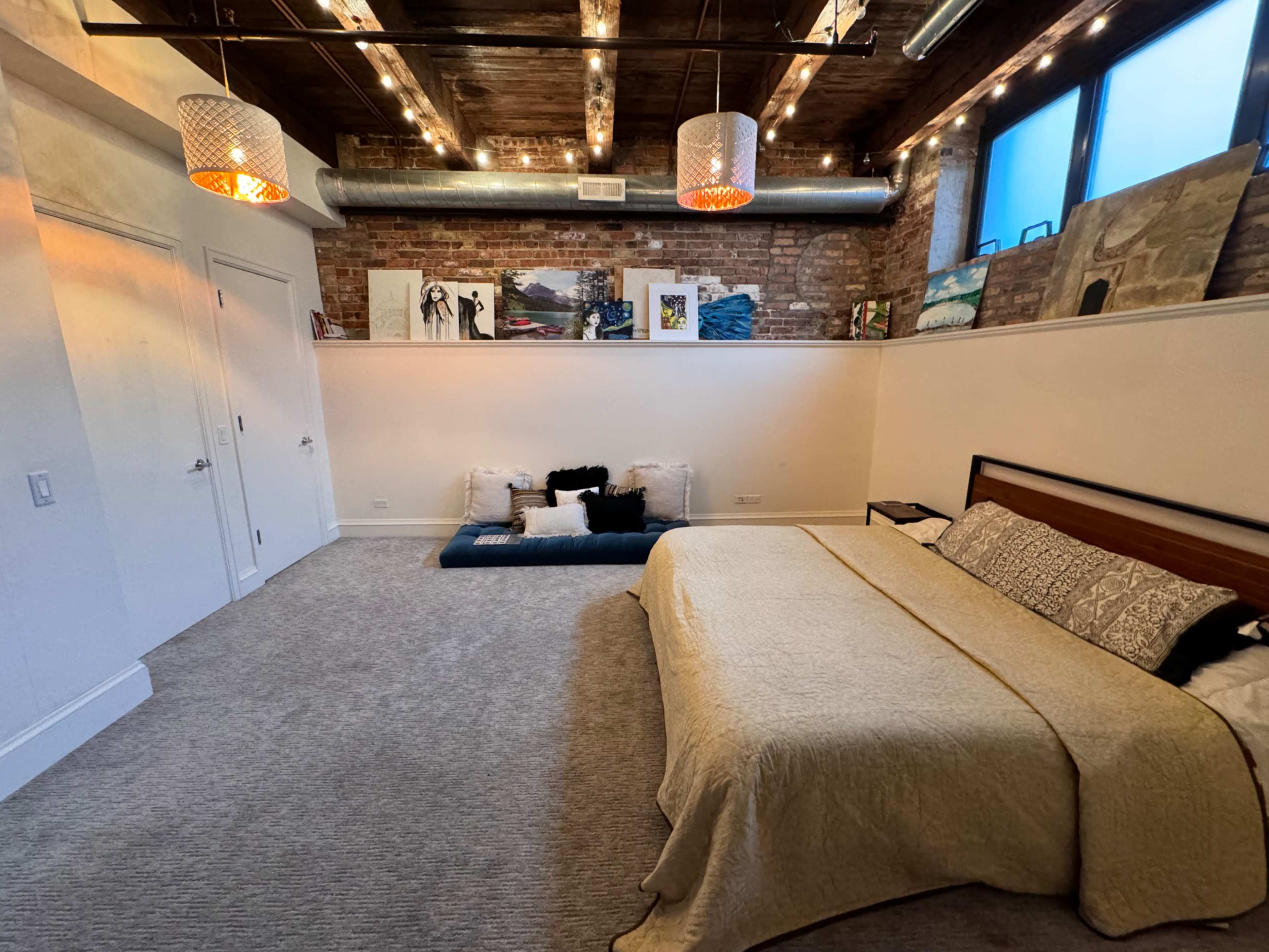 The image shows a spacious bedroom with a bed covered in a beige blanket, surrounded by decorative pillows and artwork on the walls, illuminated by hanging light fixtures.