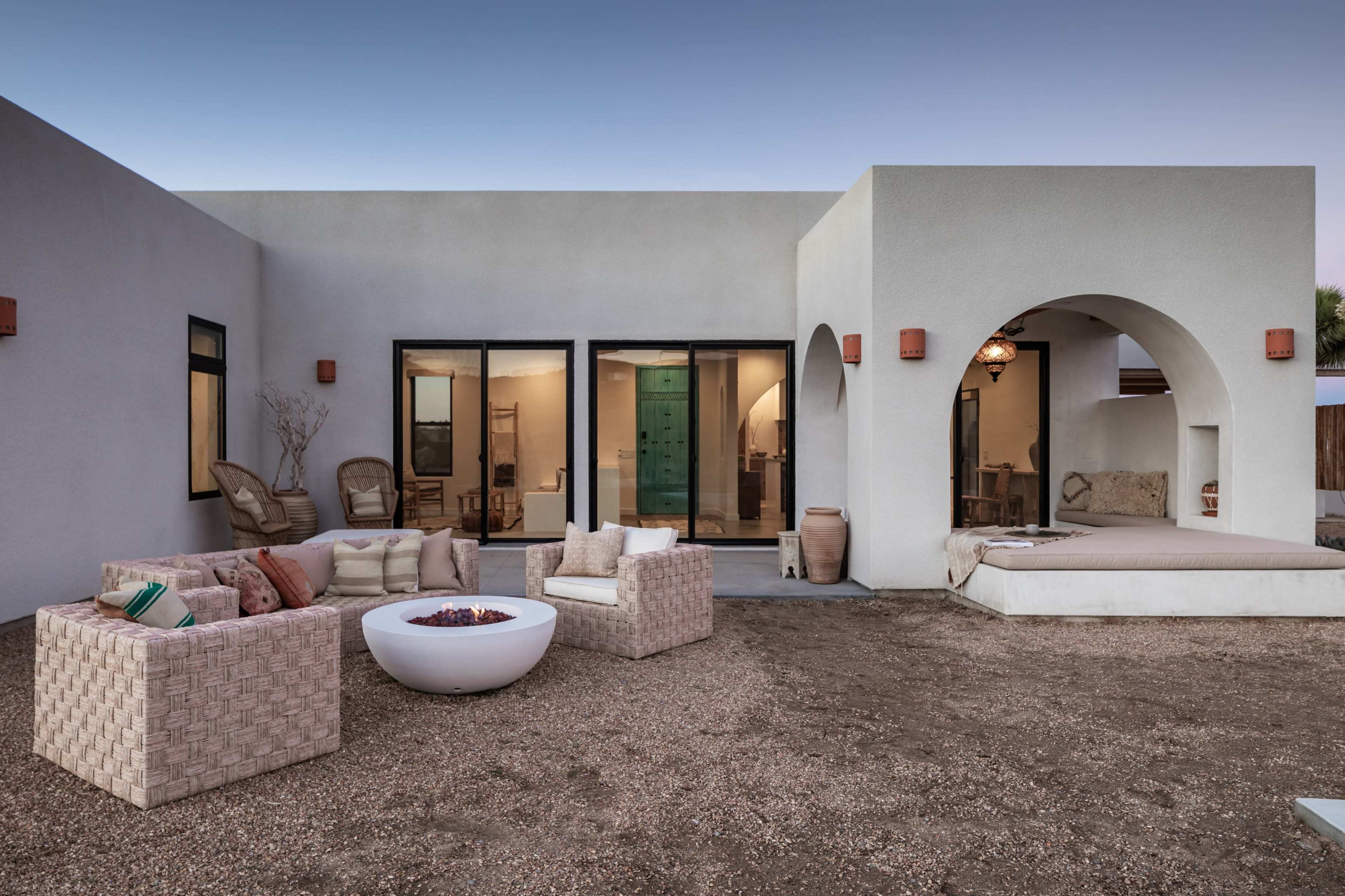 The image shows a modern, minimalist outdoor living area featuring comfortable seating, a fire pit, and large glass doors leading into a home.